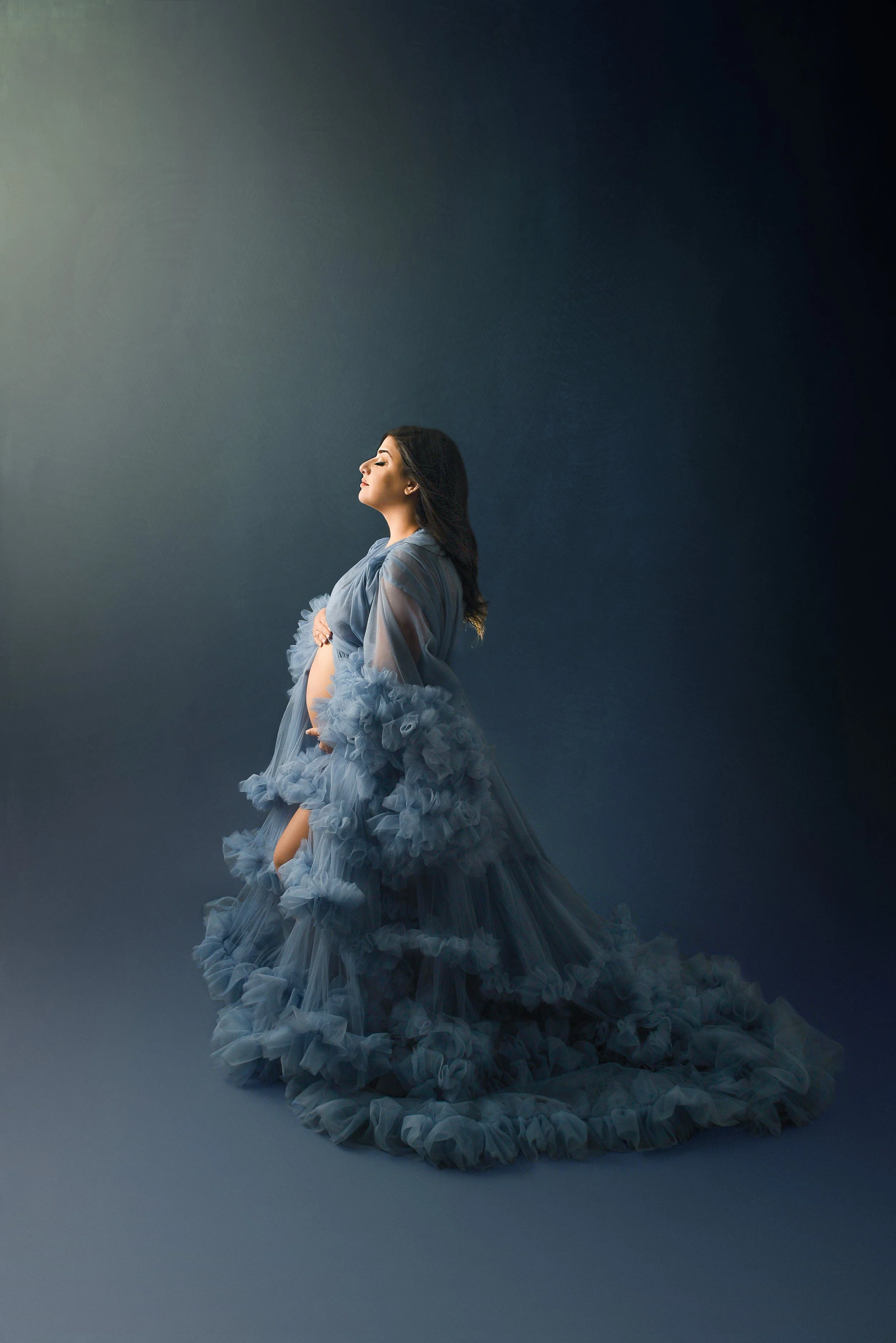 Pregnant woman in a flowing, ruffled blue gown posing with her hands on her belly against a dark, gradient background.