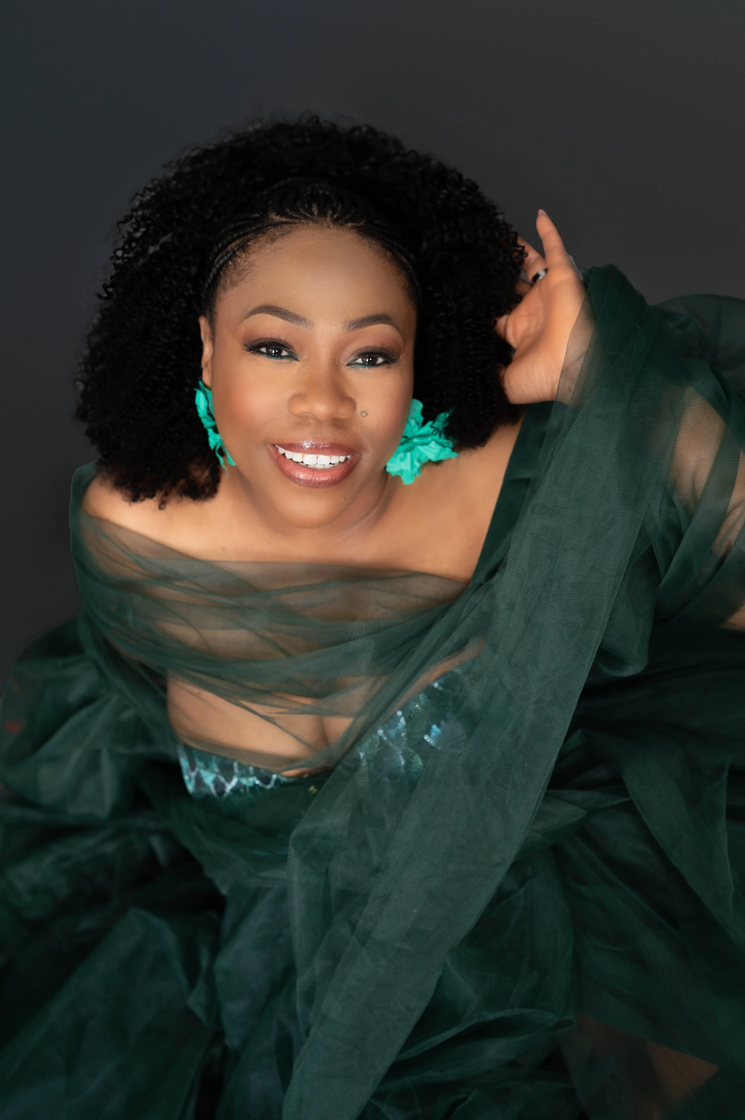 Portrait of a joyful woman wearing teal earrings and a dark green sheer dress, sitting against a dark background.