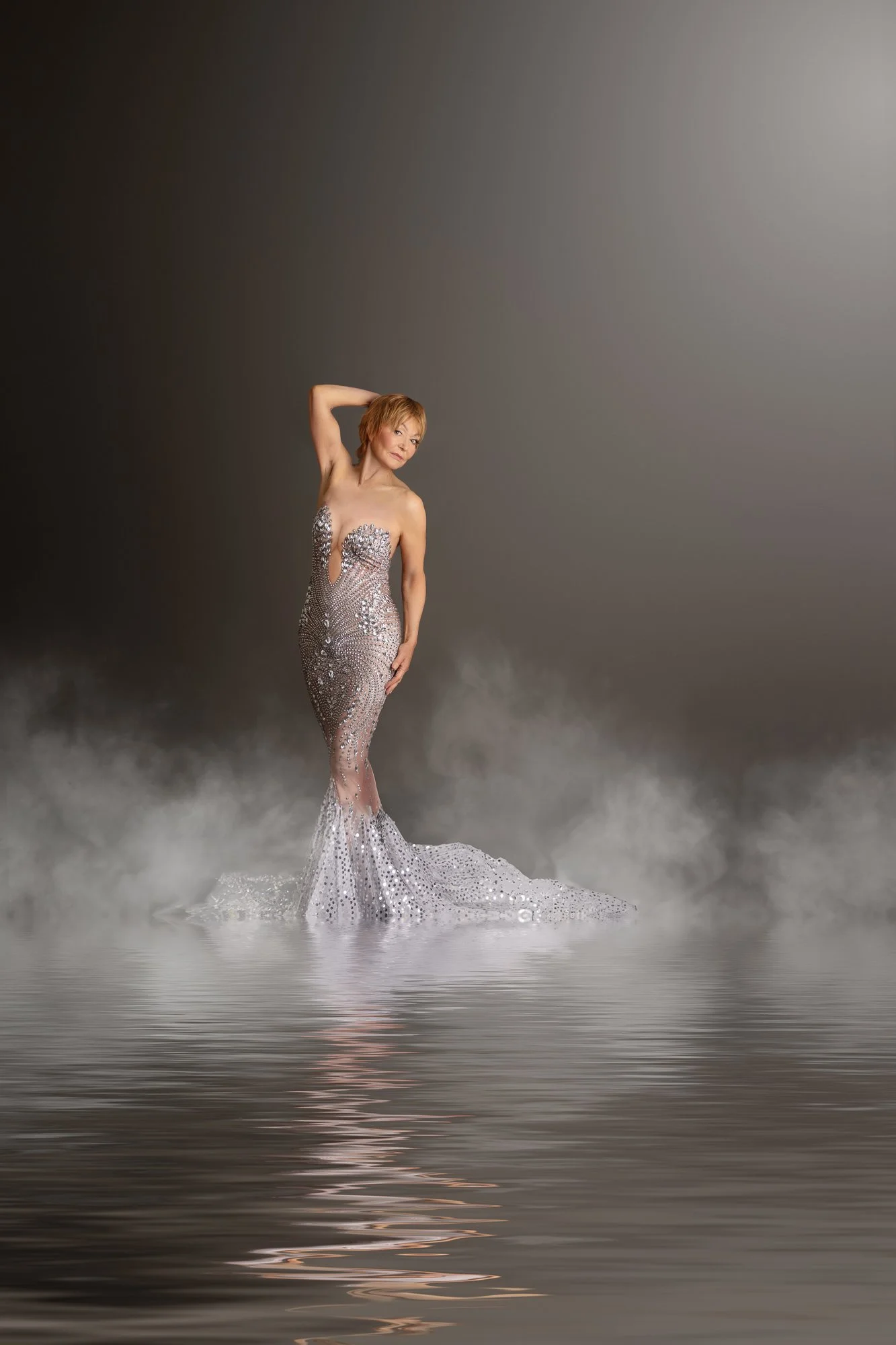 A woman in a shimmering, sequined mermaid gown standing in water with mist or fog at her feet, posing with one hand on her head and the other on her hip.
