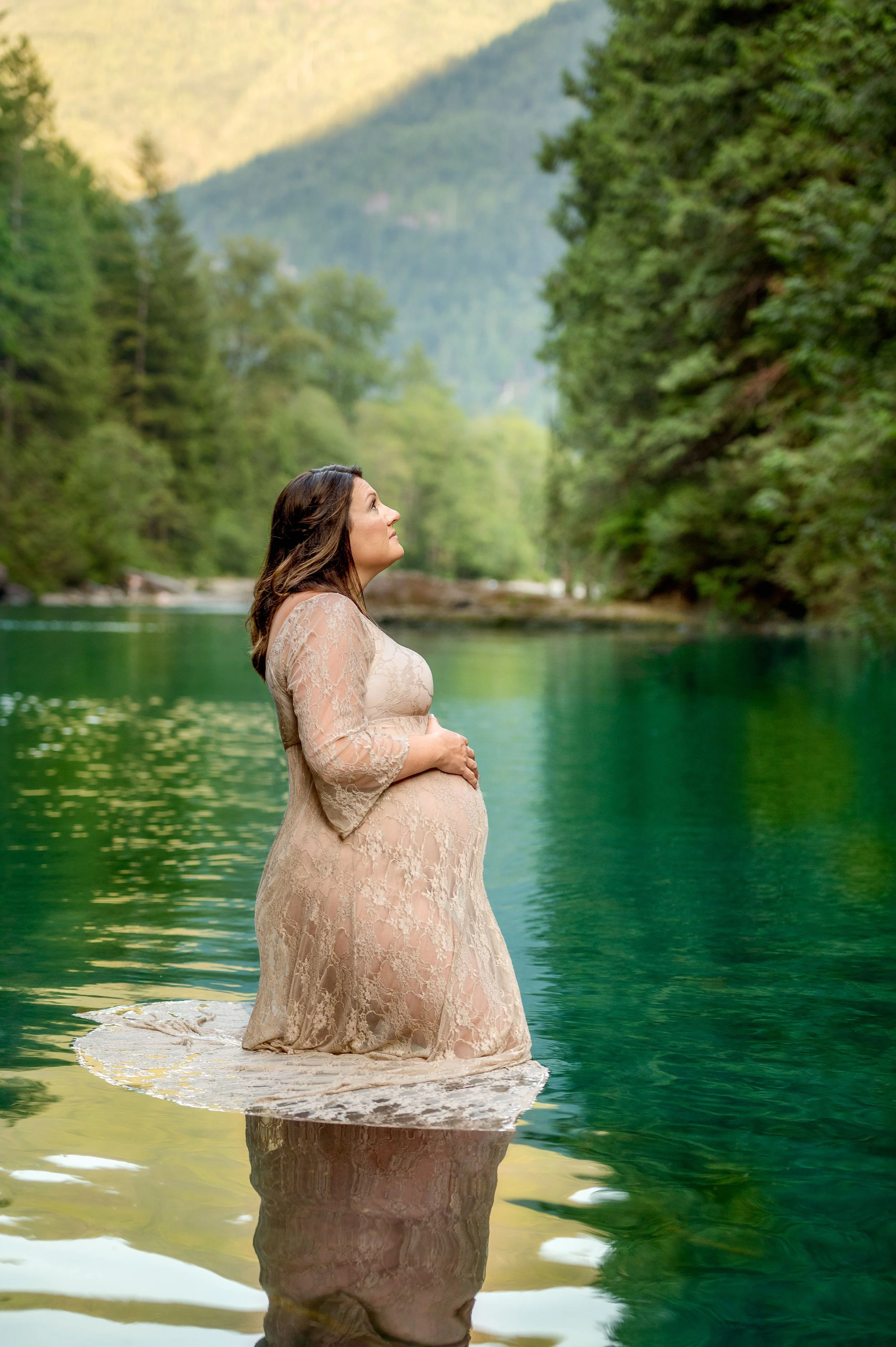 Pregnant woman kneeling in water at a river, surrounded by green trees and mountains in the background.