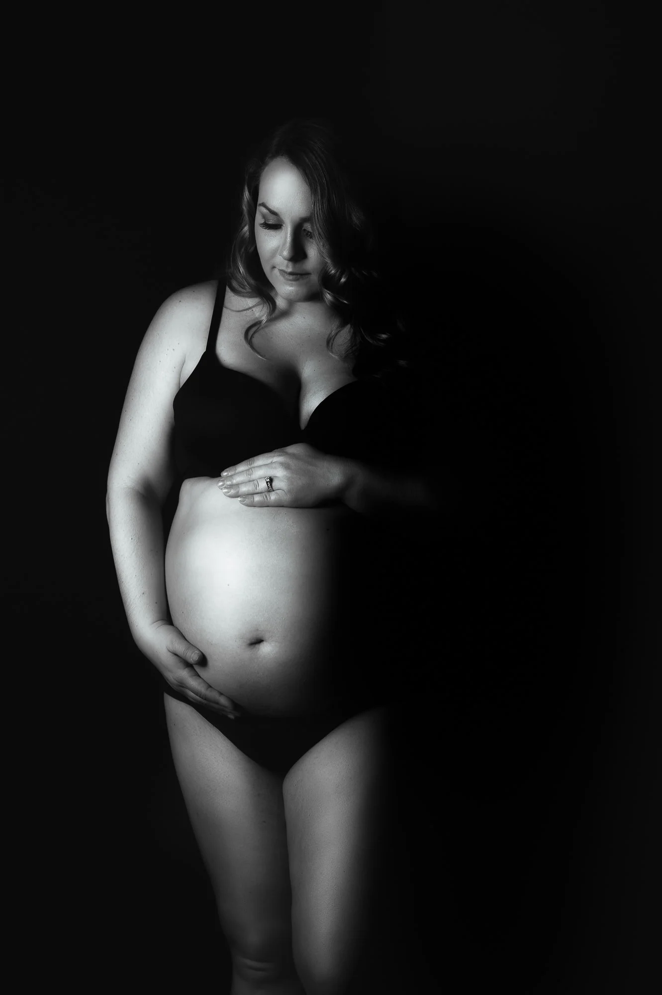 Black-and-white photograph of a pregnant woman in black lingerie, gently touching her belly, looking down thoughtfully against a dark background.