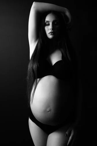 A pregnant woman with long hair posing in black underwear in a dark setting.