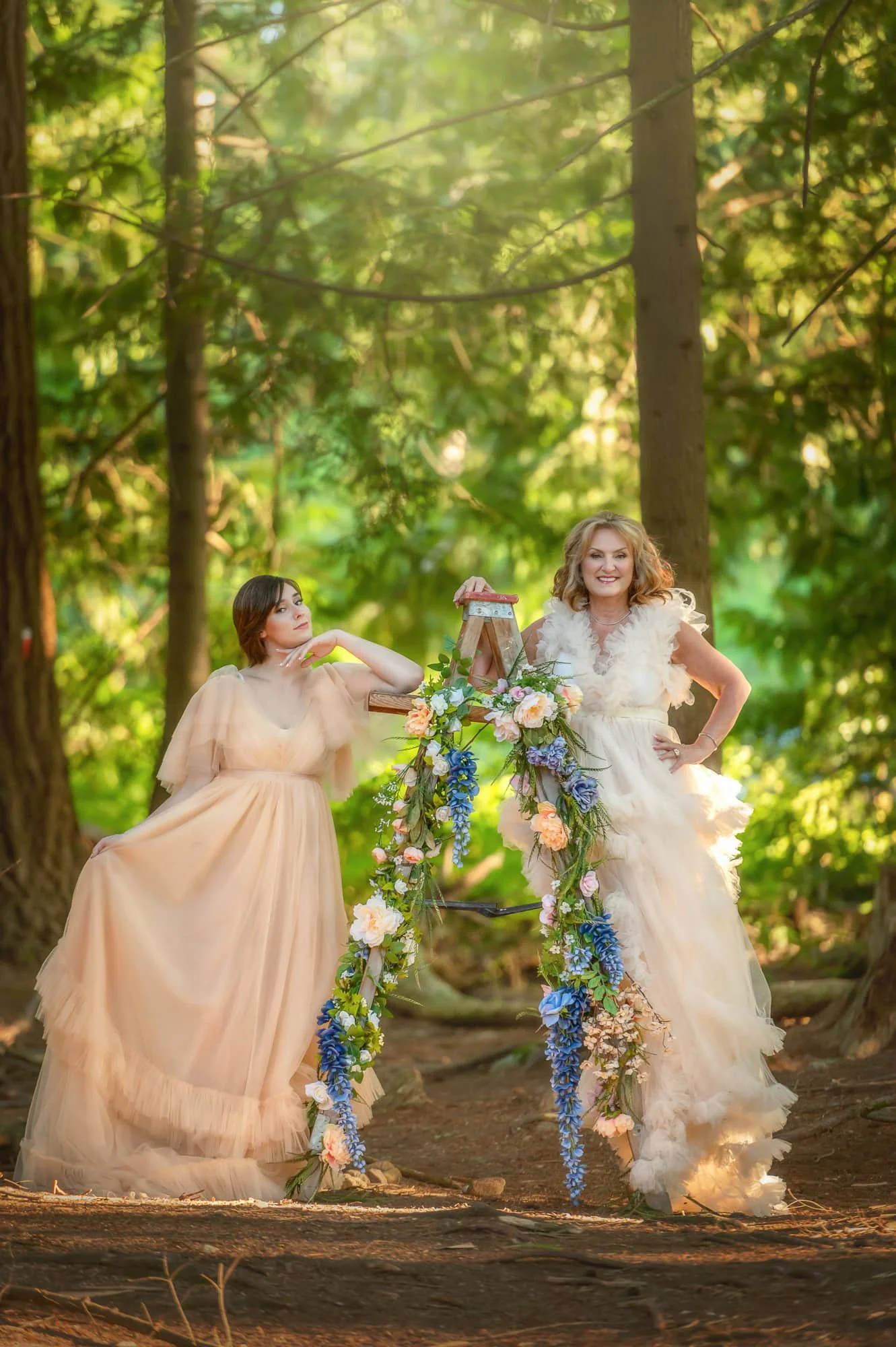 Two women in flowy dresses posing in a wooded area, with a bicycle decorated with flowers.