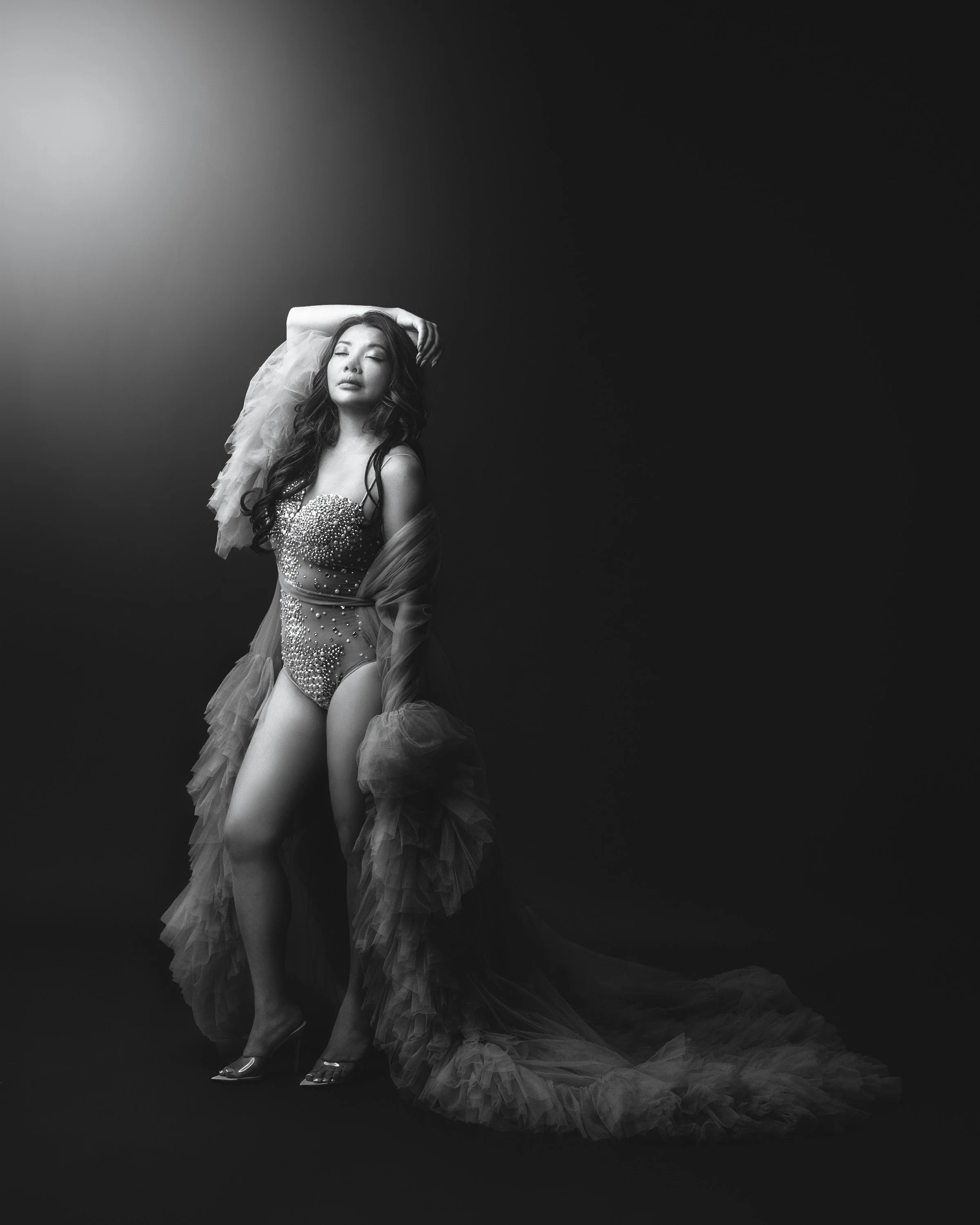 A black-and-white photo of a woman in an embellished outfit with tulle fabric, standing against a dark background with her eyes closed and one arm raised above her head.