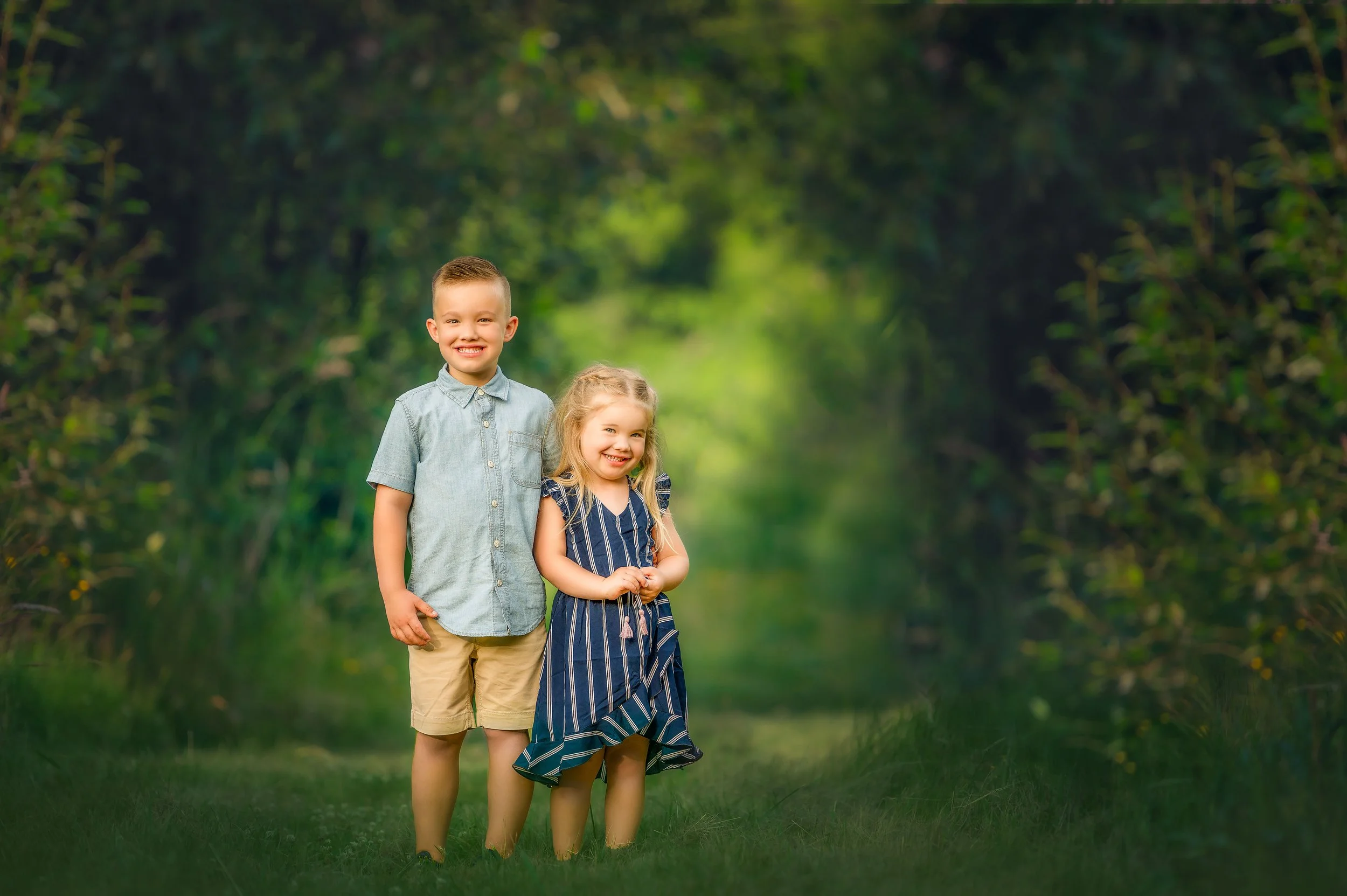 A boy and a girl standing on a grassy path in a lush green forest, smiling and looking at the camera.