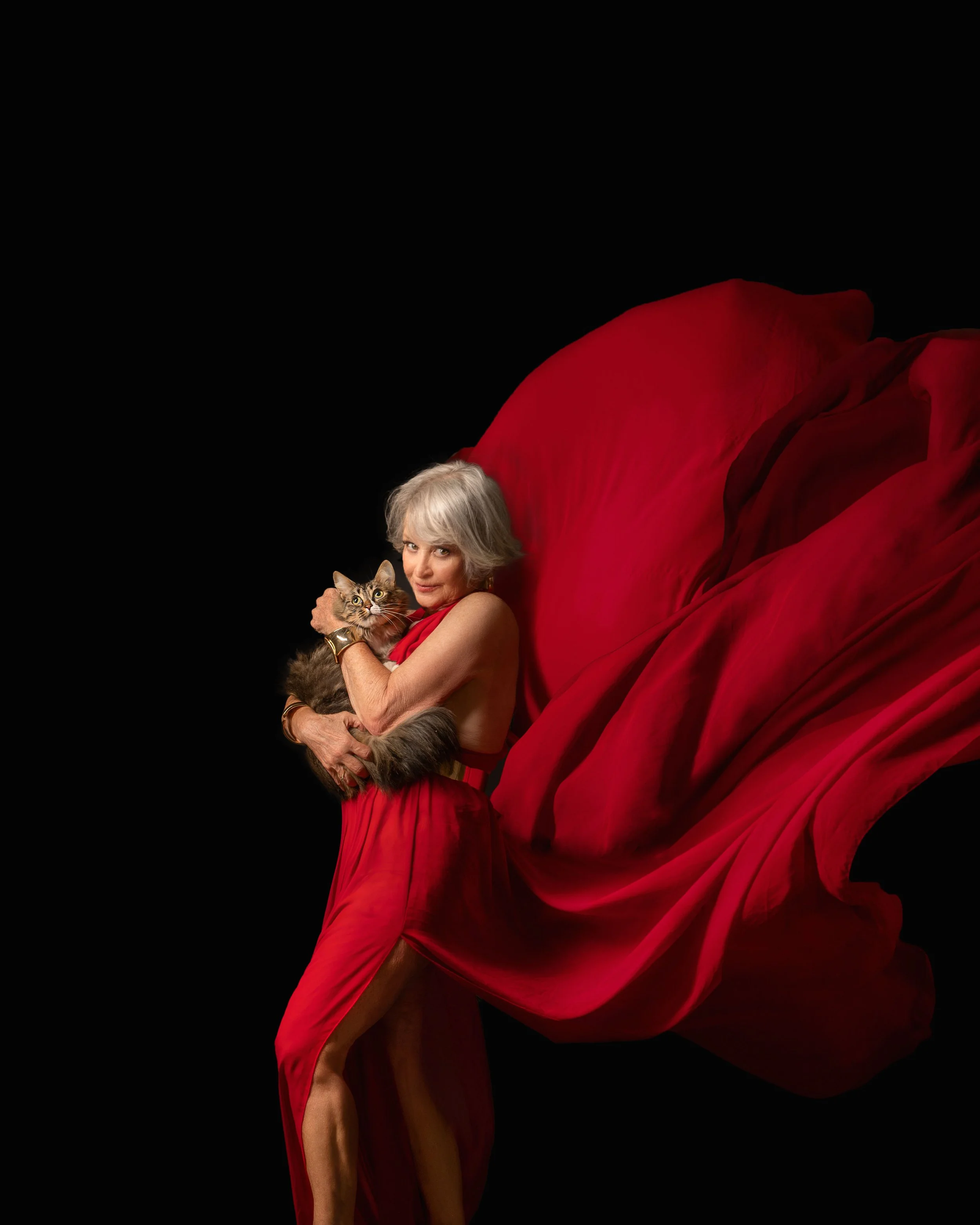 A woman with short gray hair wearing a flowing red dress holds a fluffy tabby cat against a black background.