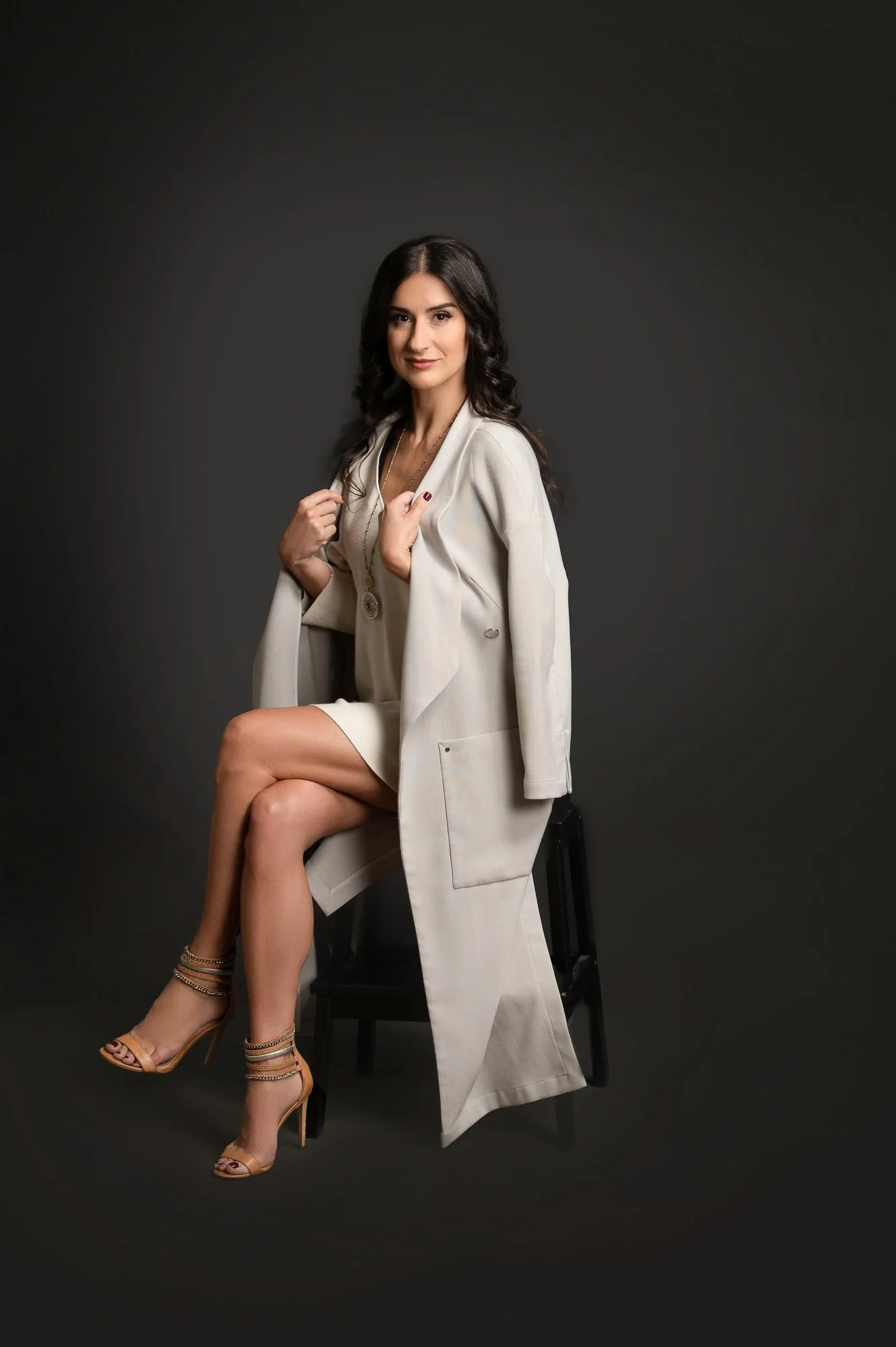 A woman with long dark hair sitting on a stool against a dark background, wearing a light-colored dress, a light coat, and high heels, looking at the camera.