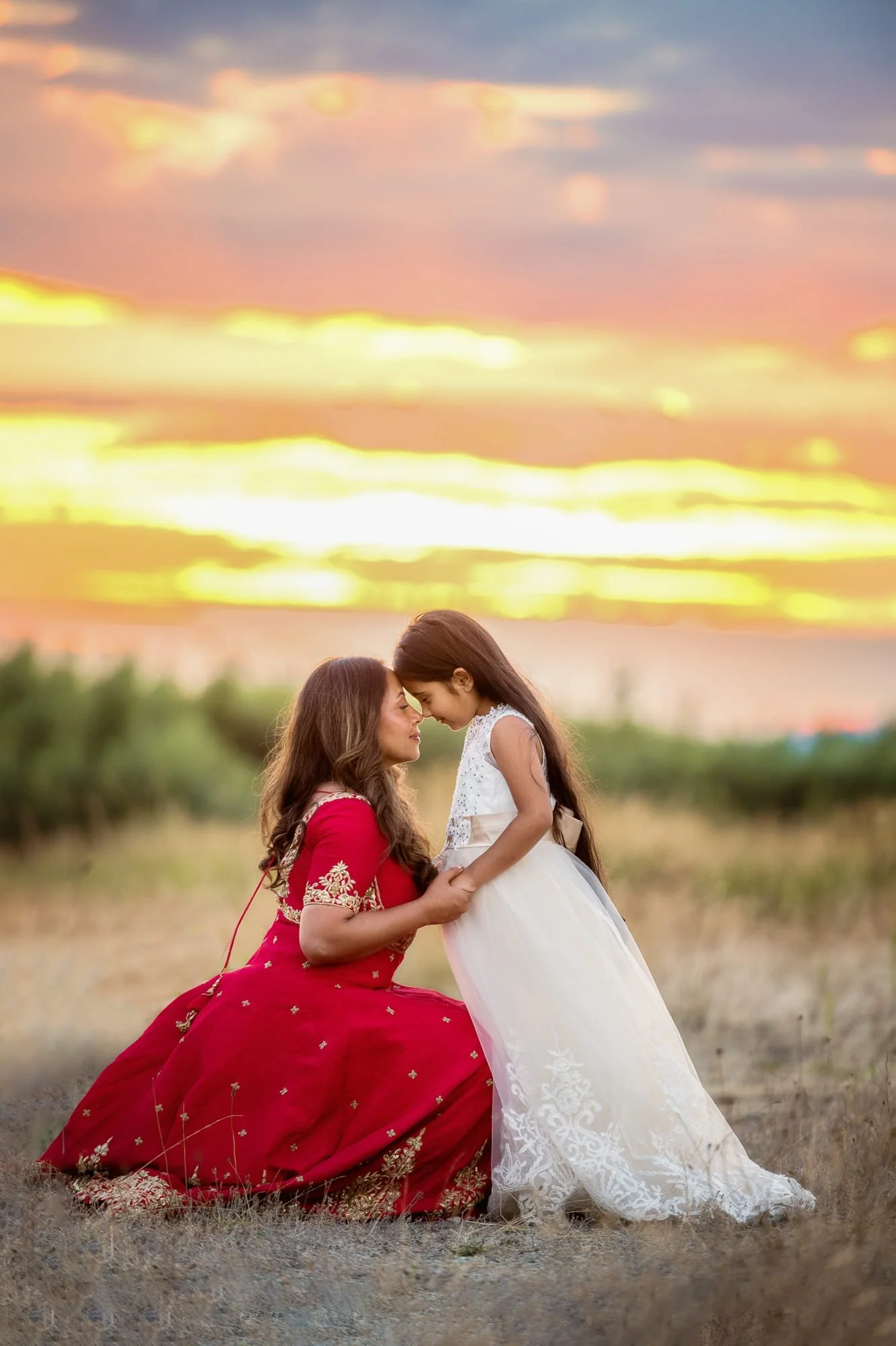 A woman in a red dress and a young girl in a white dress touching foreheads in a field during sunset.