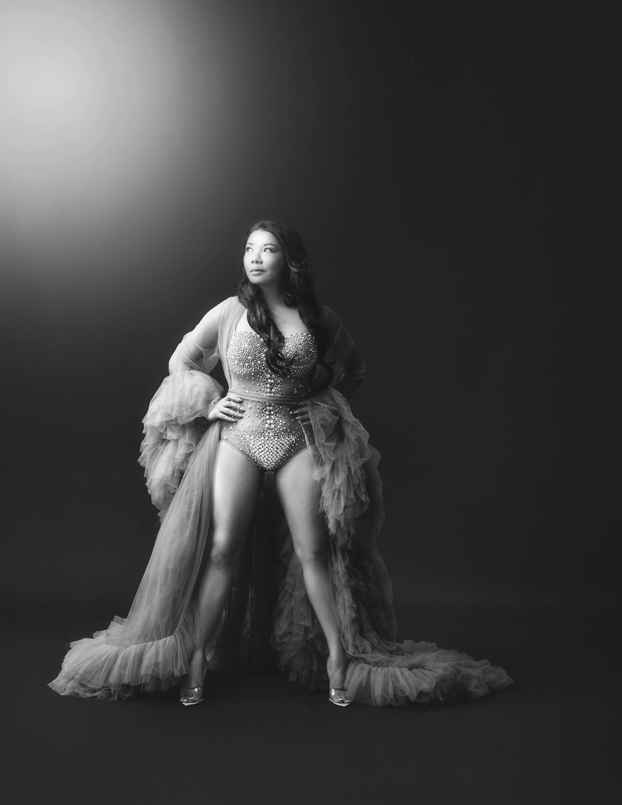 Black and white photo of a woman in a glamorous, embellished gown with ruffled tulle details, standing against a dark background with a spotlight overhead.