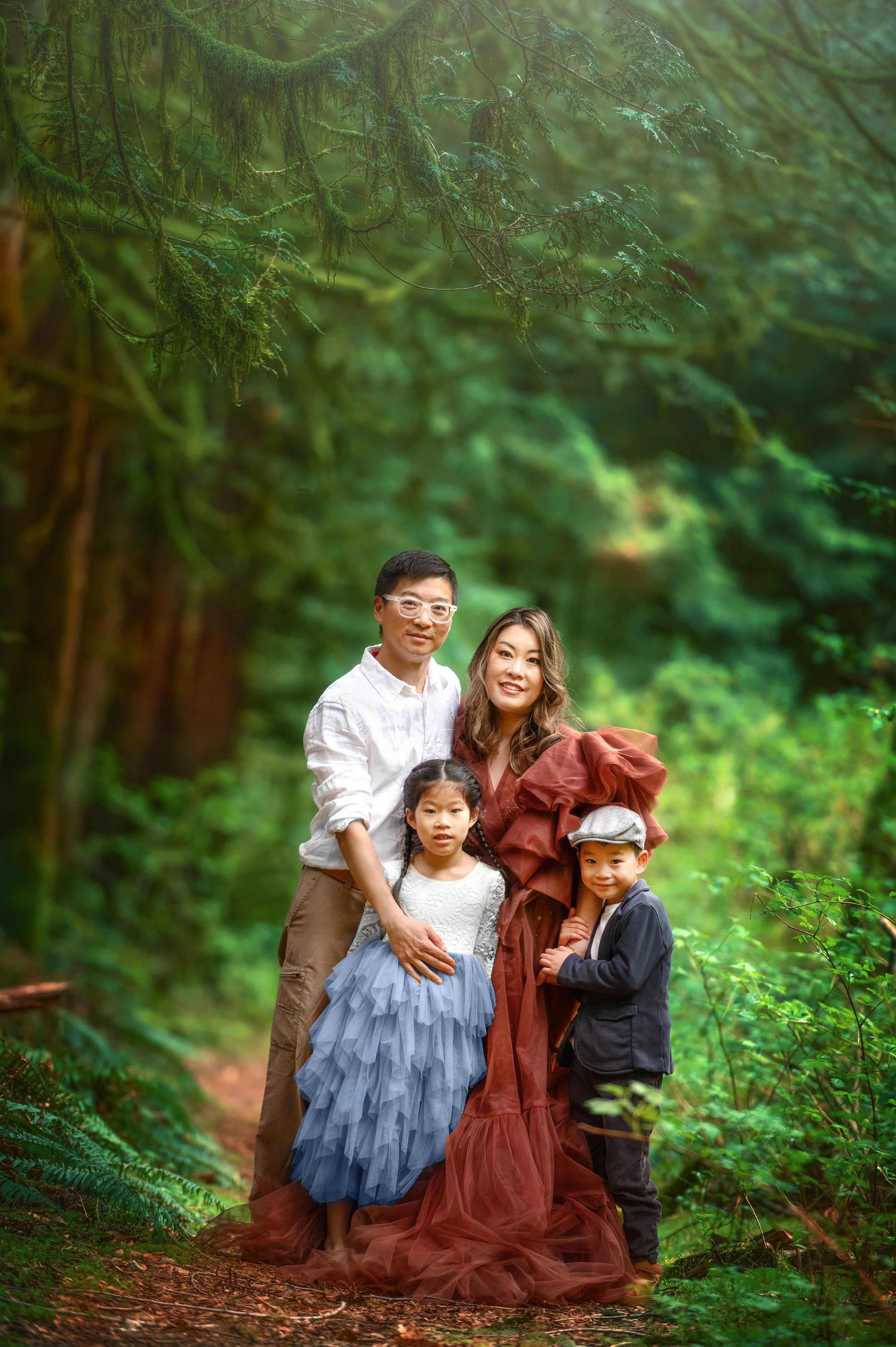 A family of four posing for a photo in a lush, green forest. The father is wearing glasses and a white shirt. The mother is dressed in a large, flowing, rust-colored gown. Their daughter is in a fancy, tiered, light blue dress, and their son is weari