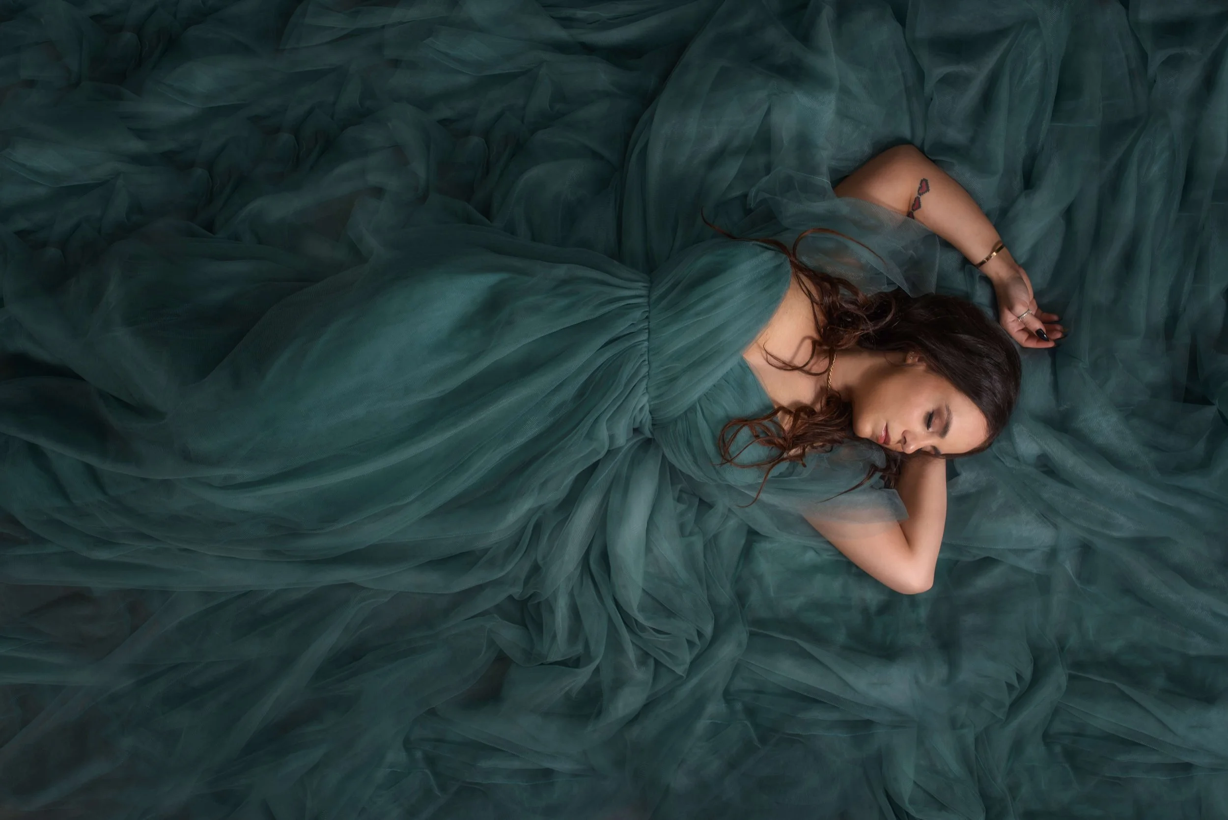 A woman in a flowing emerald green dress lying on a bed of matching fabric, with her eyes closed and her head resting on her arm.