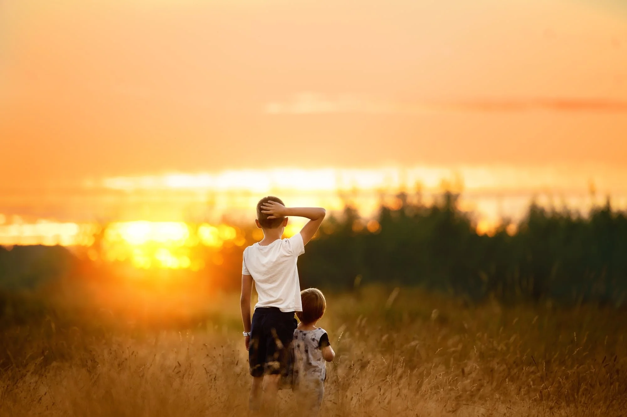 A boy and a girl standing in a grassy field during sunset, with the boy shielding his eyes and looking into the distance.