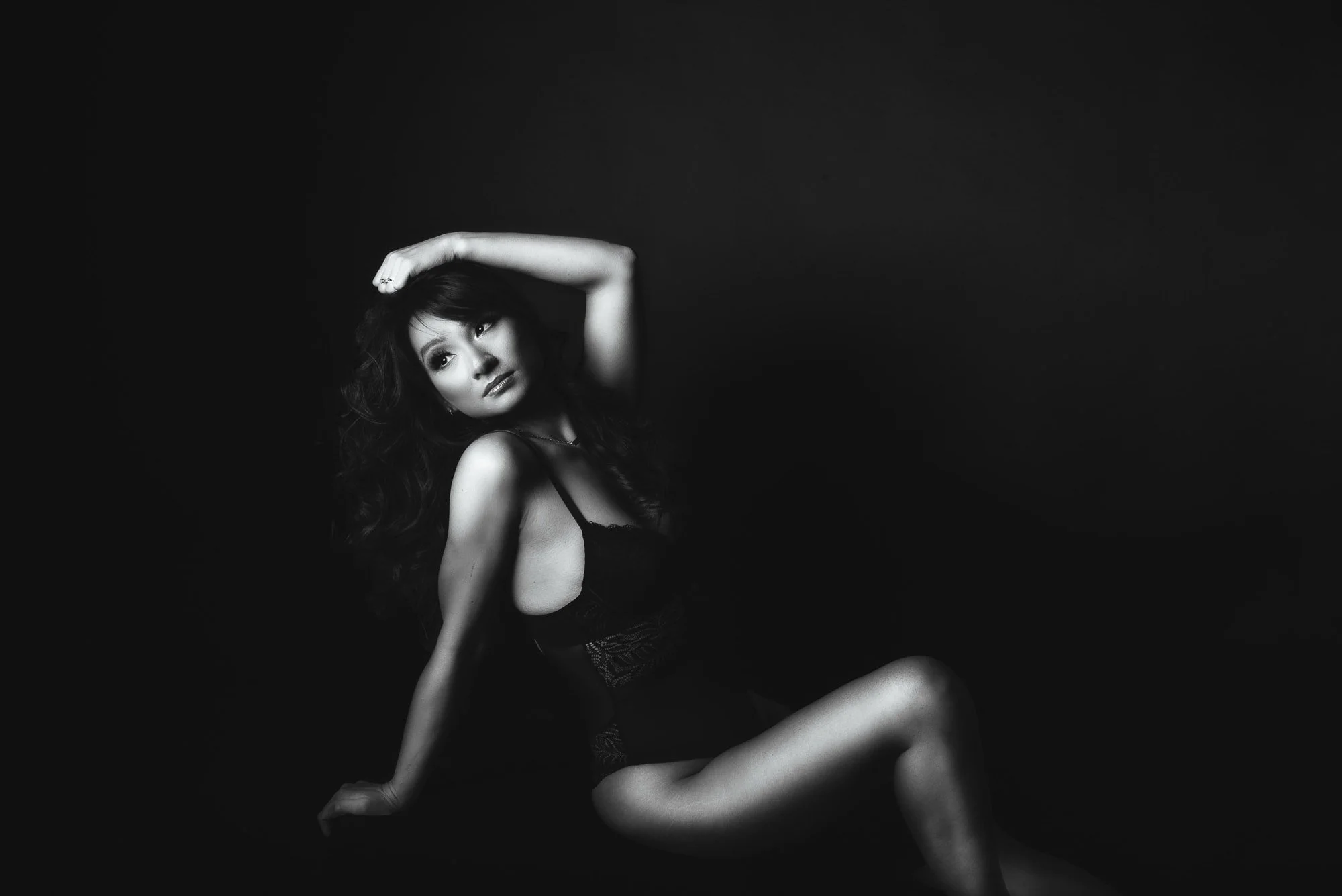 A woman with long dark hair, wearing black lingerie, sitting against a black background, with one arm raised and her hand touching her head.