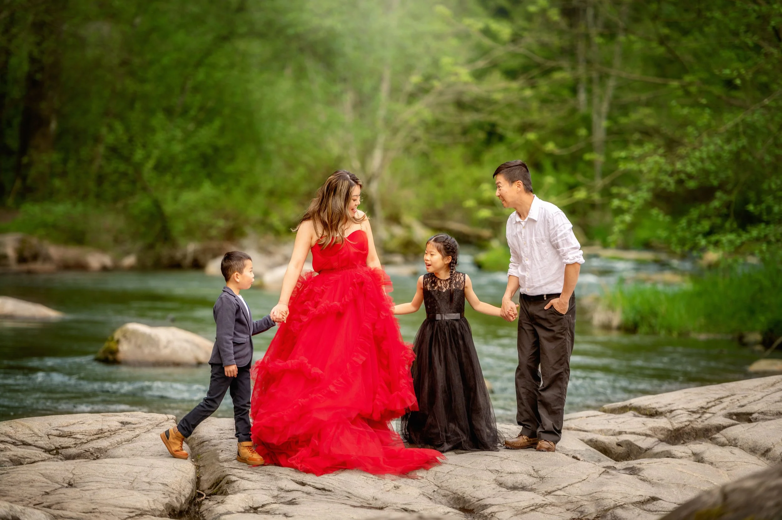A family of four, dressed elegantly, stands on rocks near a river in a forest. The mother wears a red gown, the father a white shirt and dark pants, the young girl in a black dress, and the young boy in a dark jacket. They are smiling and holding han