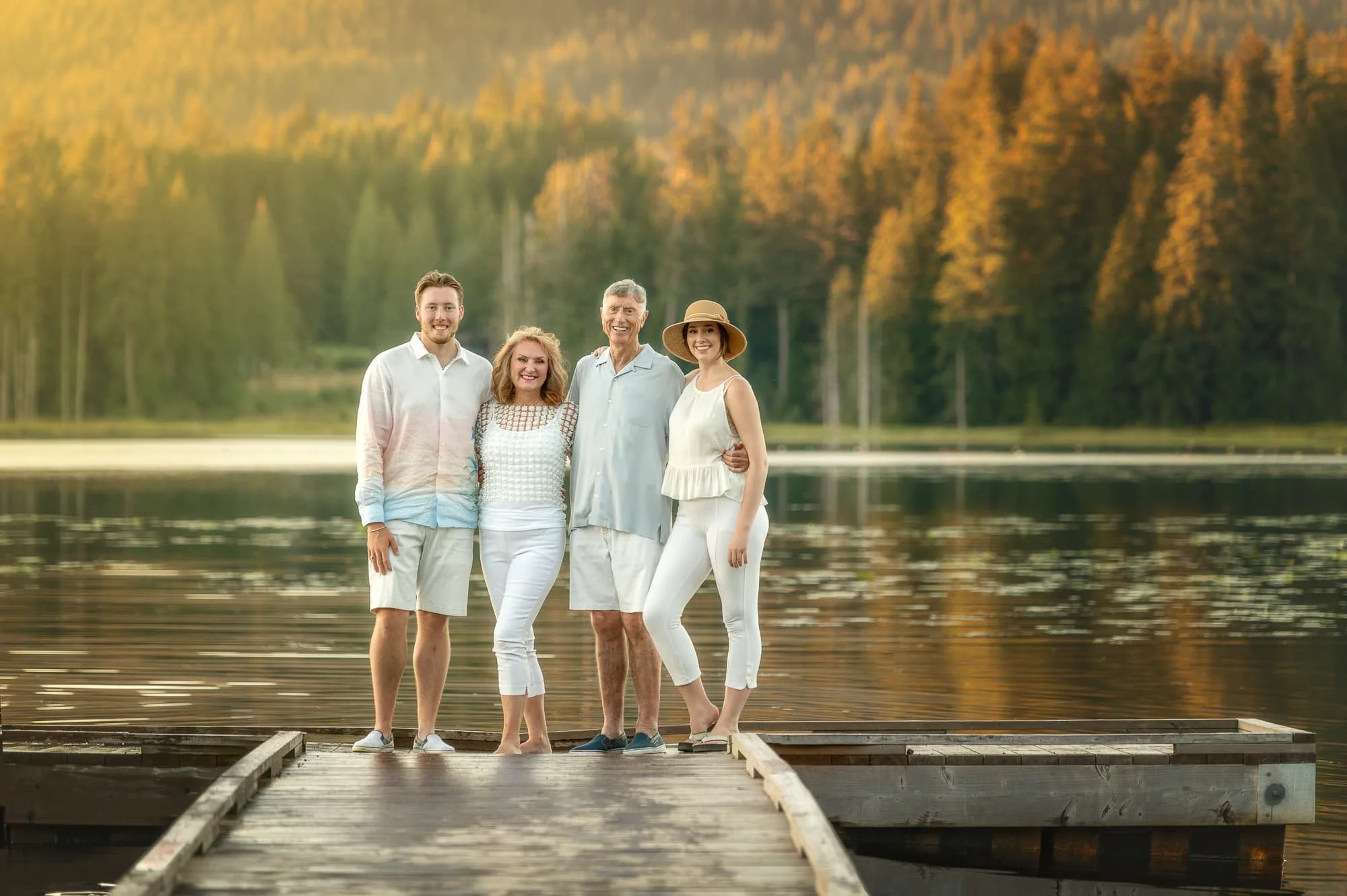 Family of four standing on a dock by a lake, smiling with trees in the background during sunset.