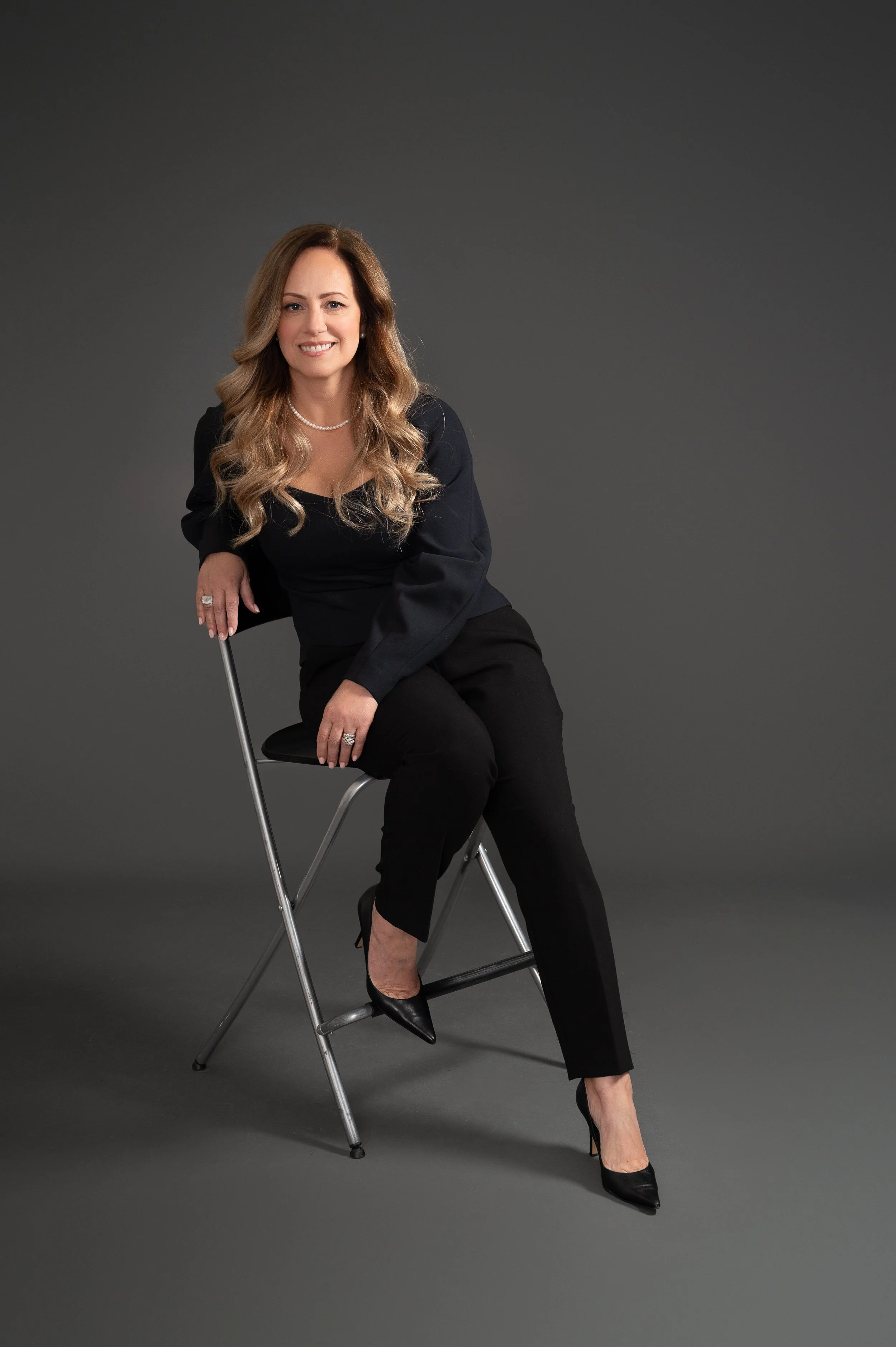 A woman with long, wavy blonde hair, dressed in a black outfit with black high heels, sitting on a black chair with a metal frame, smiling against a dark gray background.