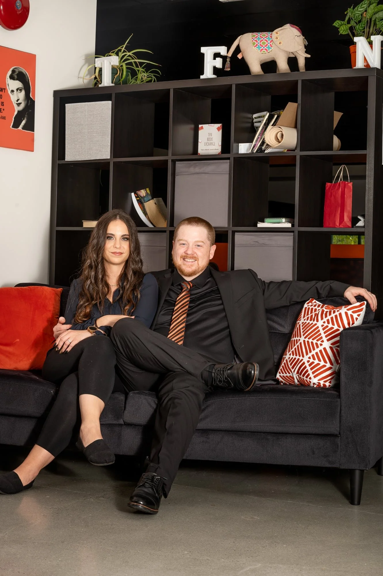 A man and woman sitting on a black sofa in front of a bookshelf with various books and decorations. The man is wearing a black suit with a striped tie, and the woman is wearing a dark blue blouse and black pants. The sofa has red and white pillows. T