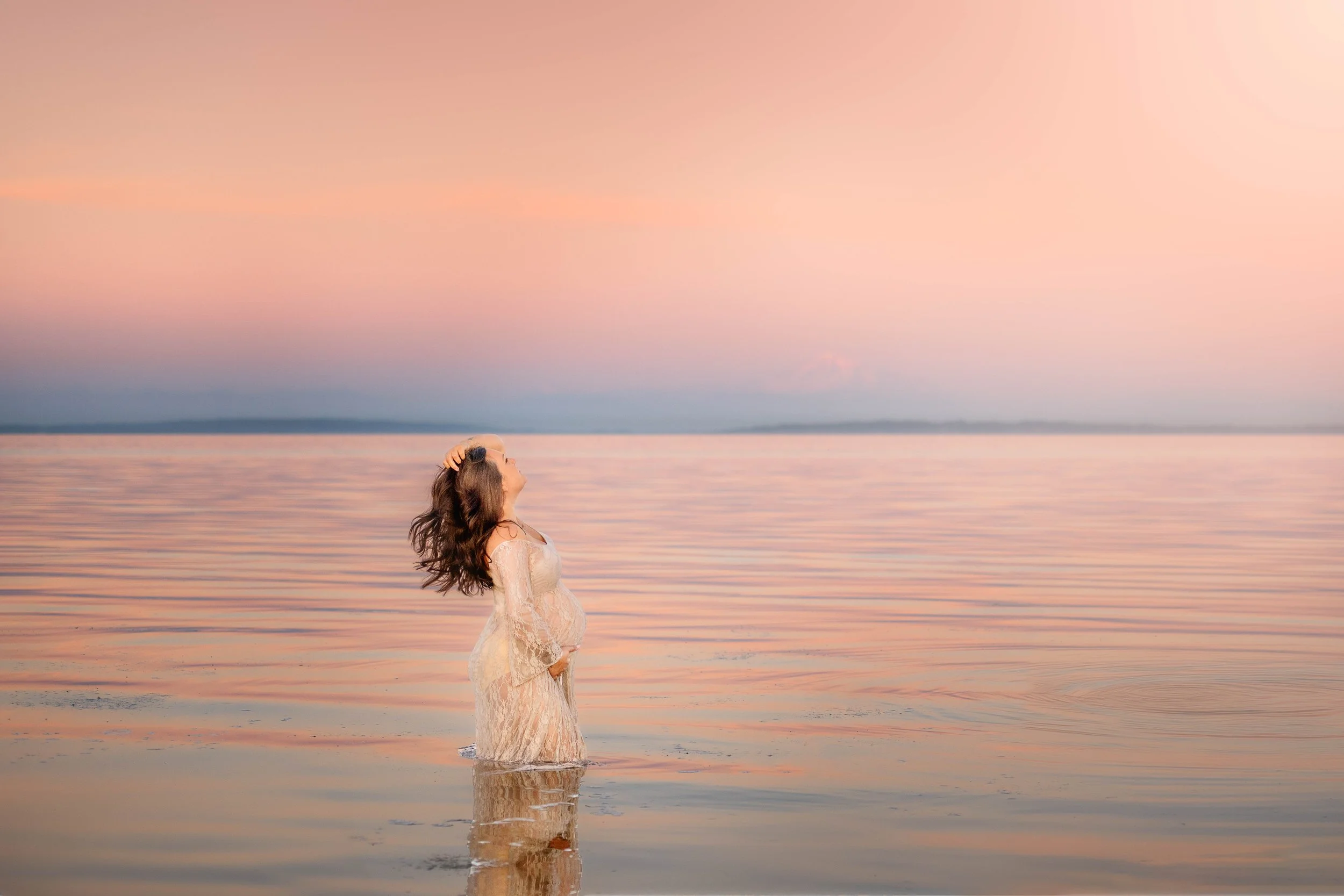 A woman in a white lace dress standing in shallow water during sunset or sunrise, with her head tilted back and one hand on her hair.