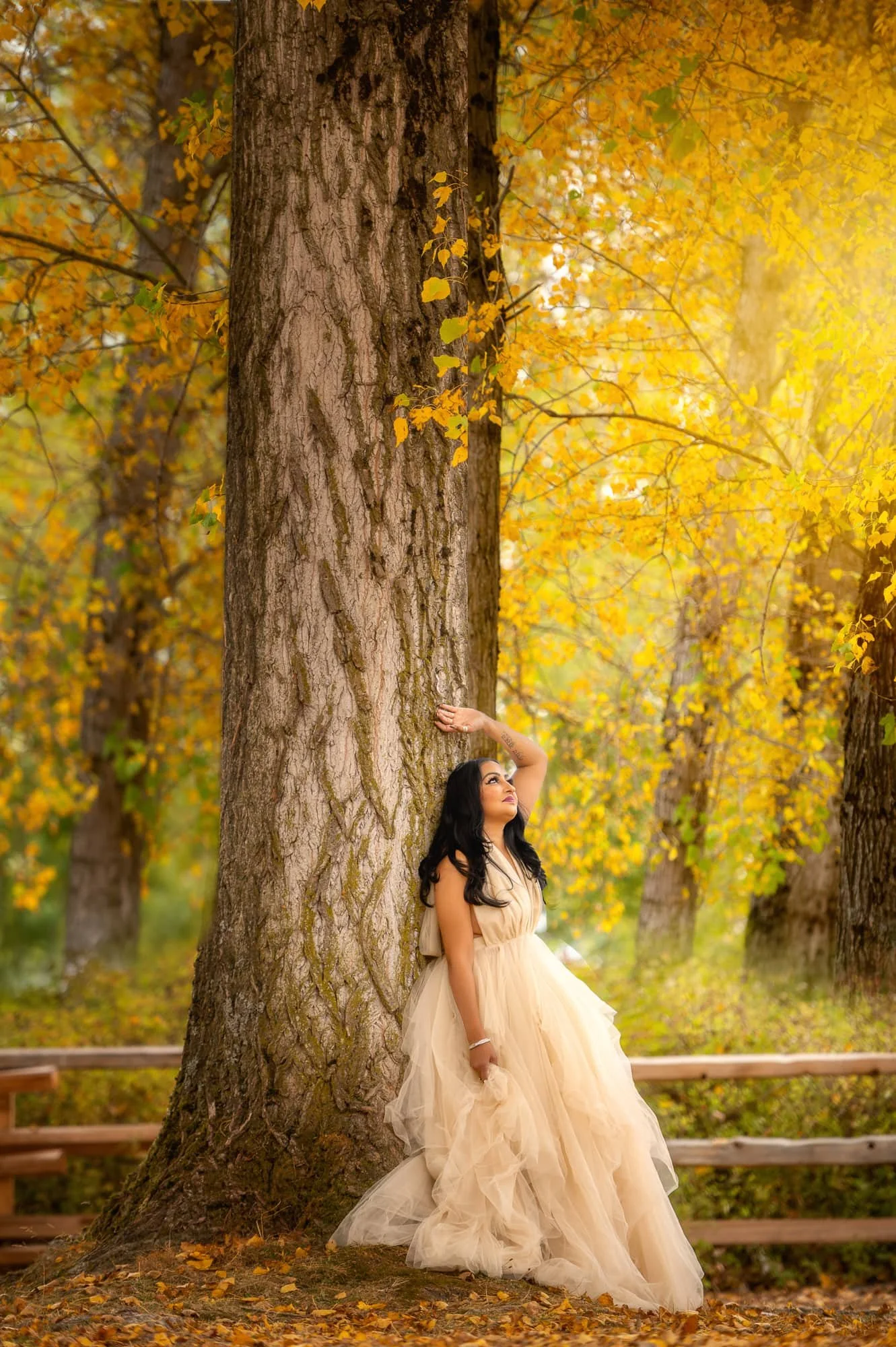 A woman in a cream-colored gown leaning against a large tree in a forest with yellow and orange autumn leaves.