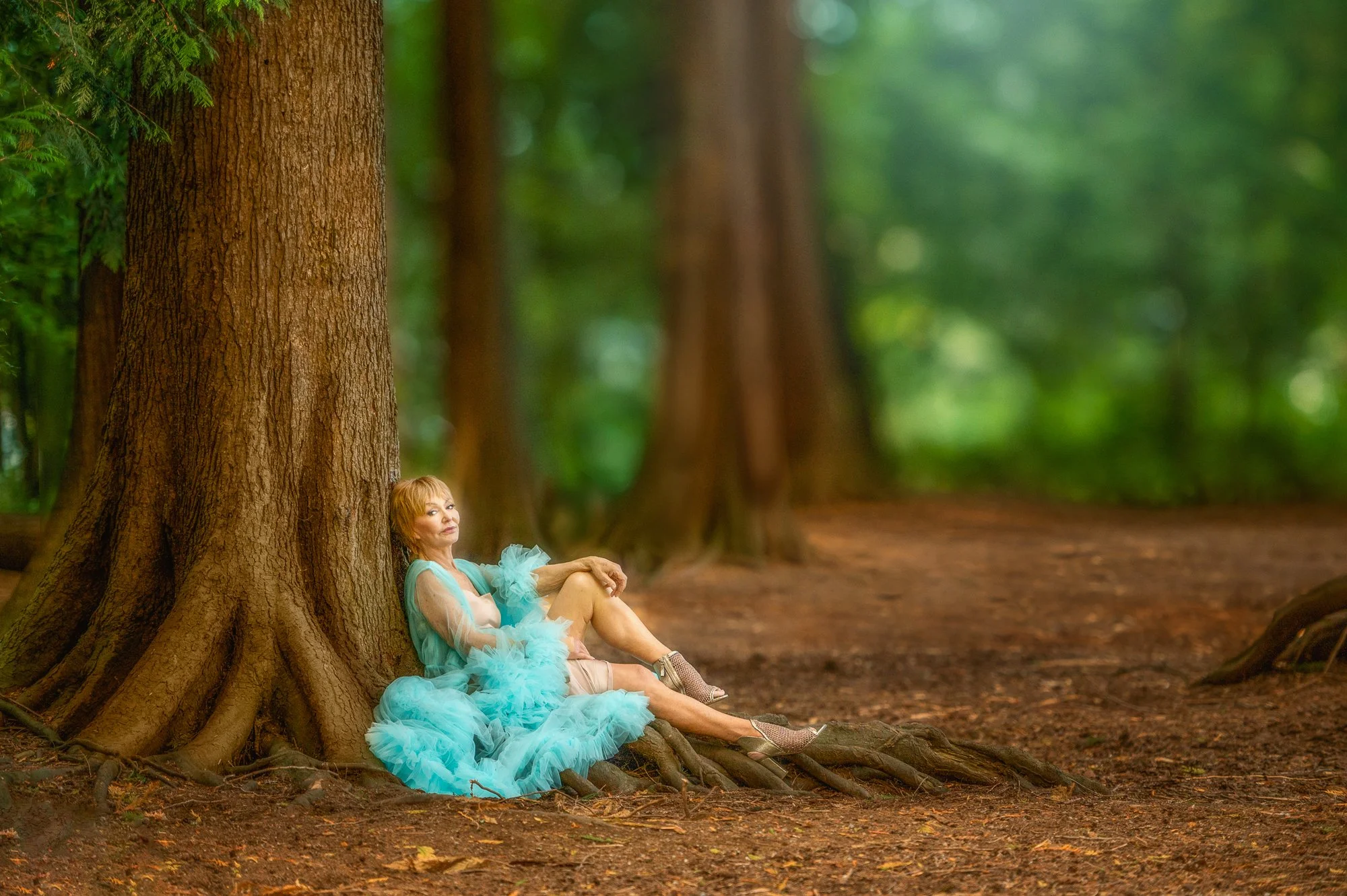 A woman in a light blue, ruffled dress and high heels sitting on tree roots at the base of a large tree in a green forest.