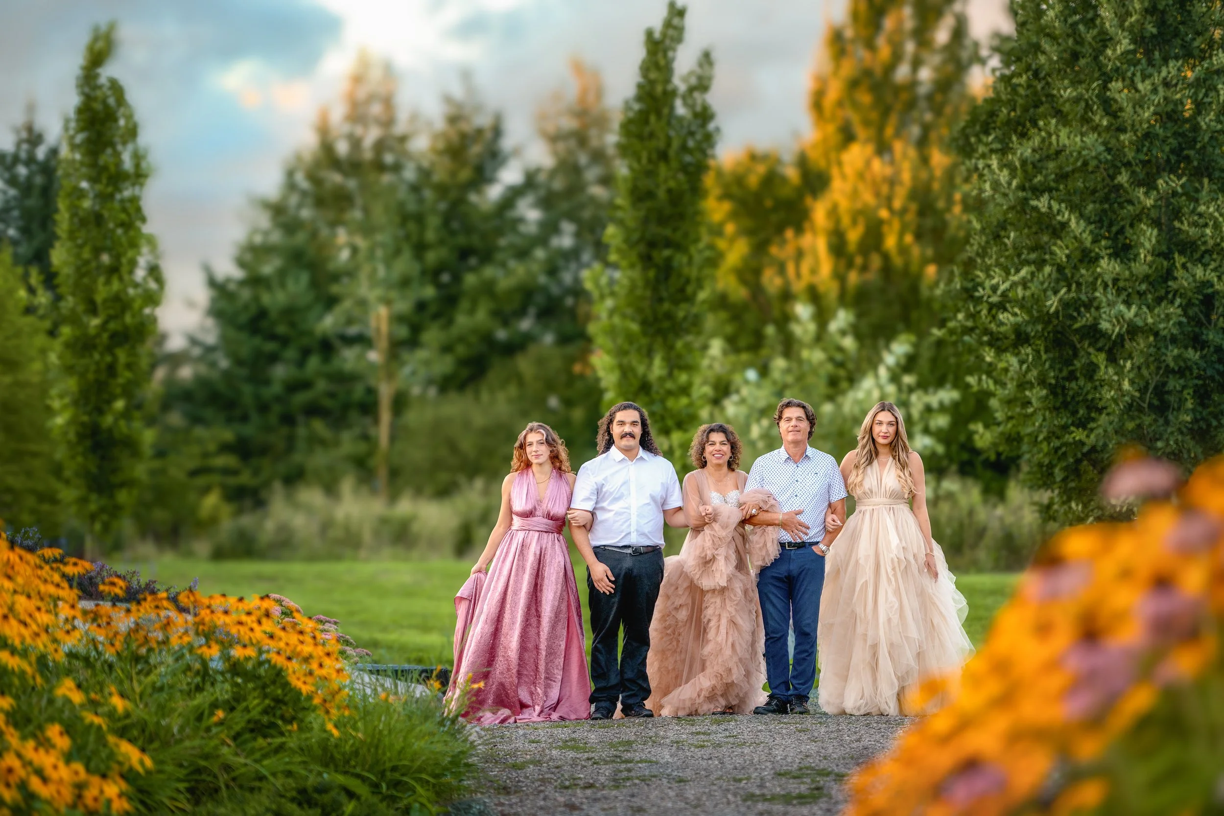A group of five people walking arm in arm outdoors during sunset, dressed in formal attire, with lush green trees and colorful flowers in the background.