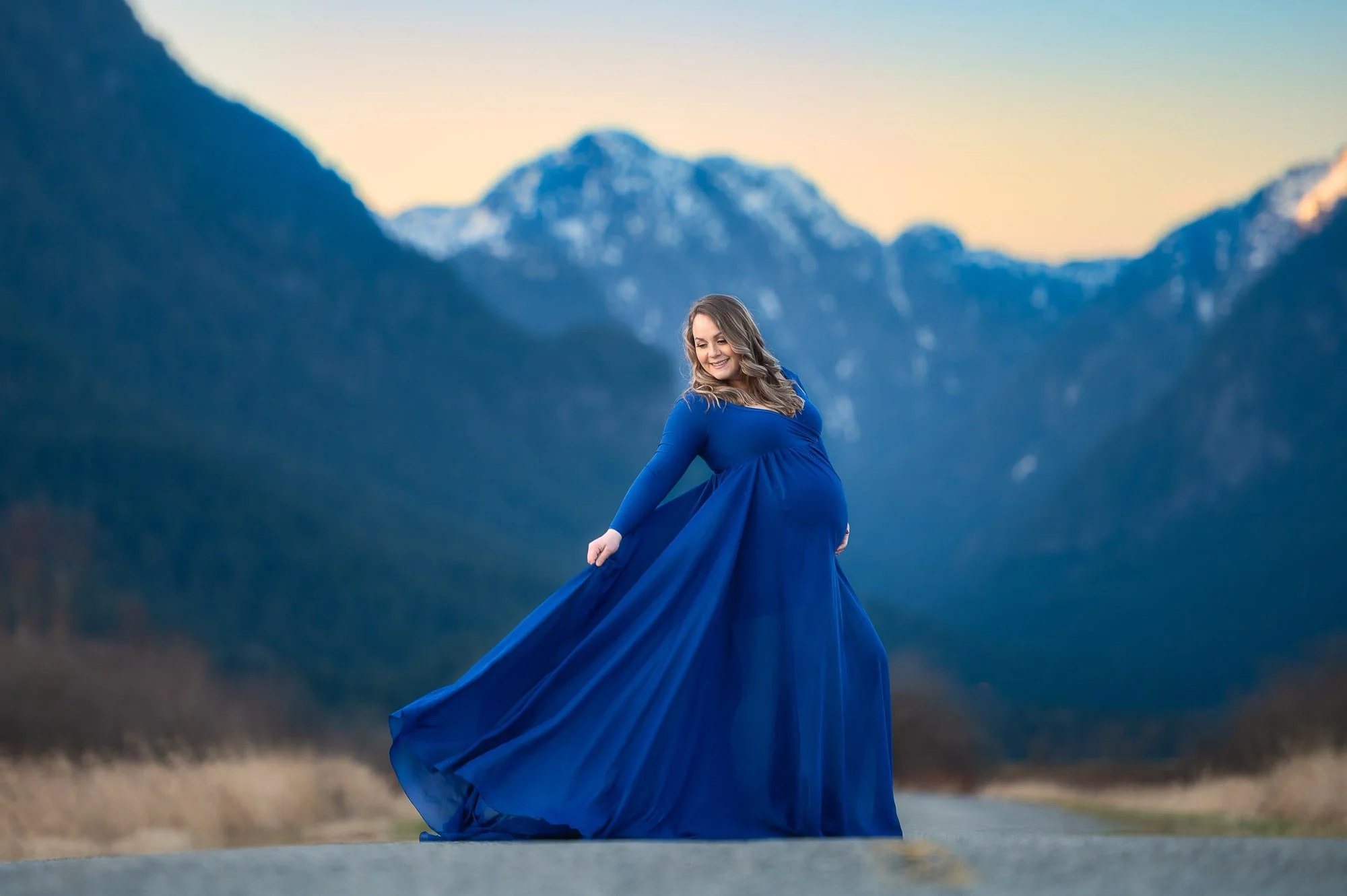 A pregnant woman in a long blue dress standing outdoors on a road, with mountainous landscape in the background during sunset.