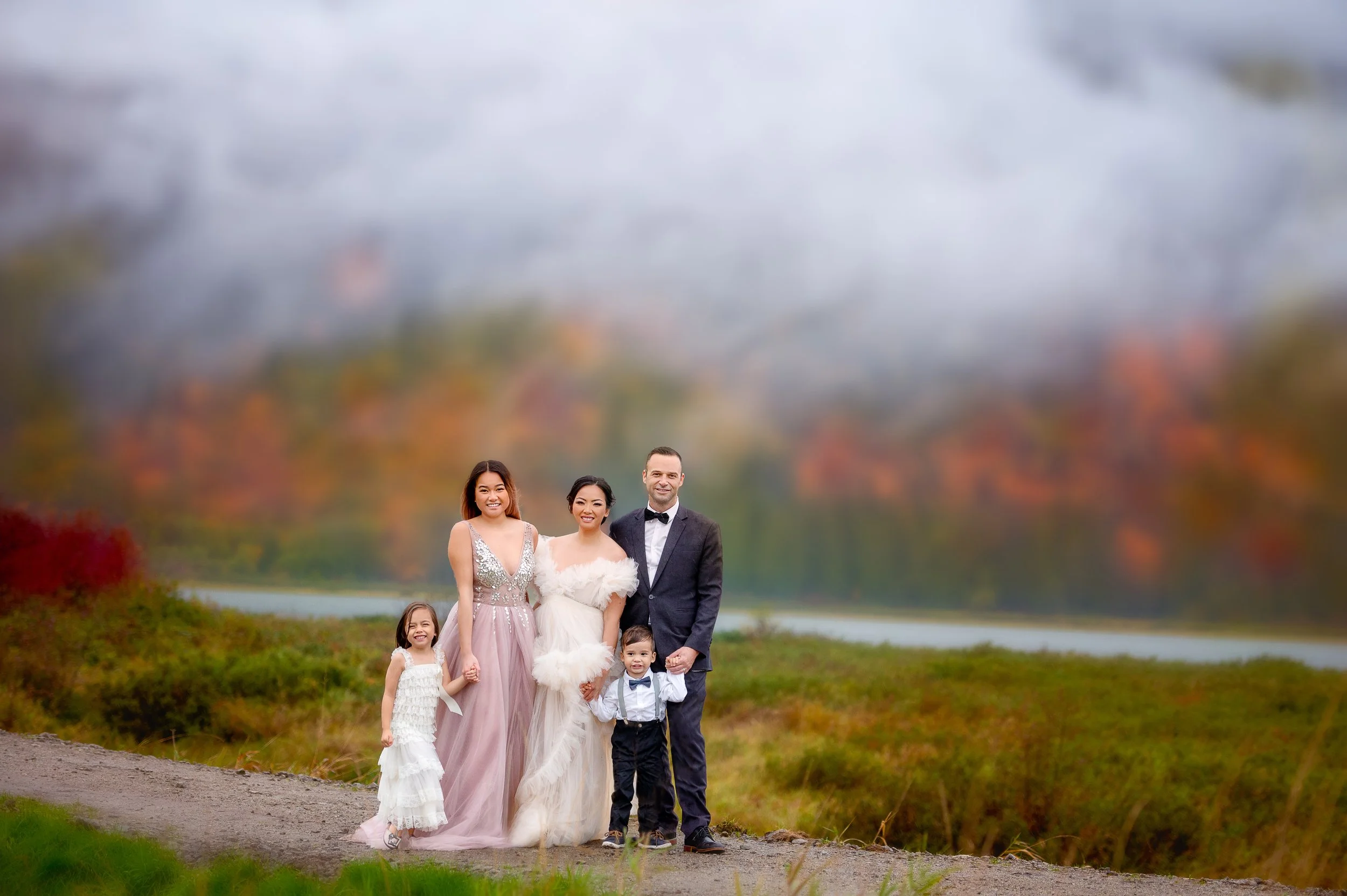 A wedding party with a bride, groom, two women, and two children standing outdoors on a path with trees and hills in the background, some of which are colorful autumn foliage.
