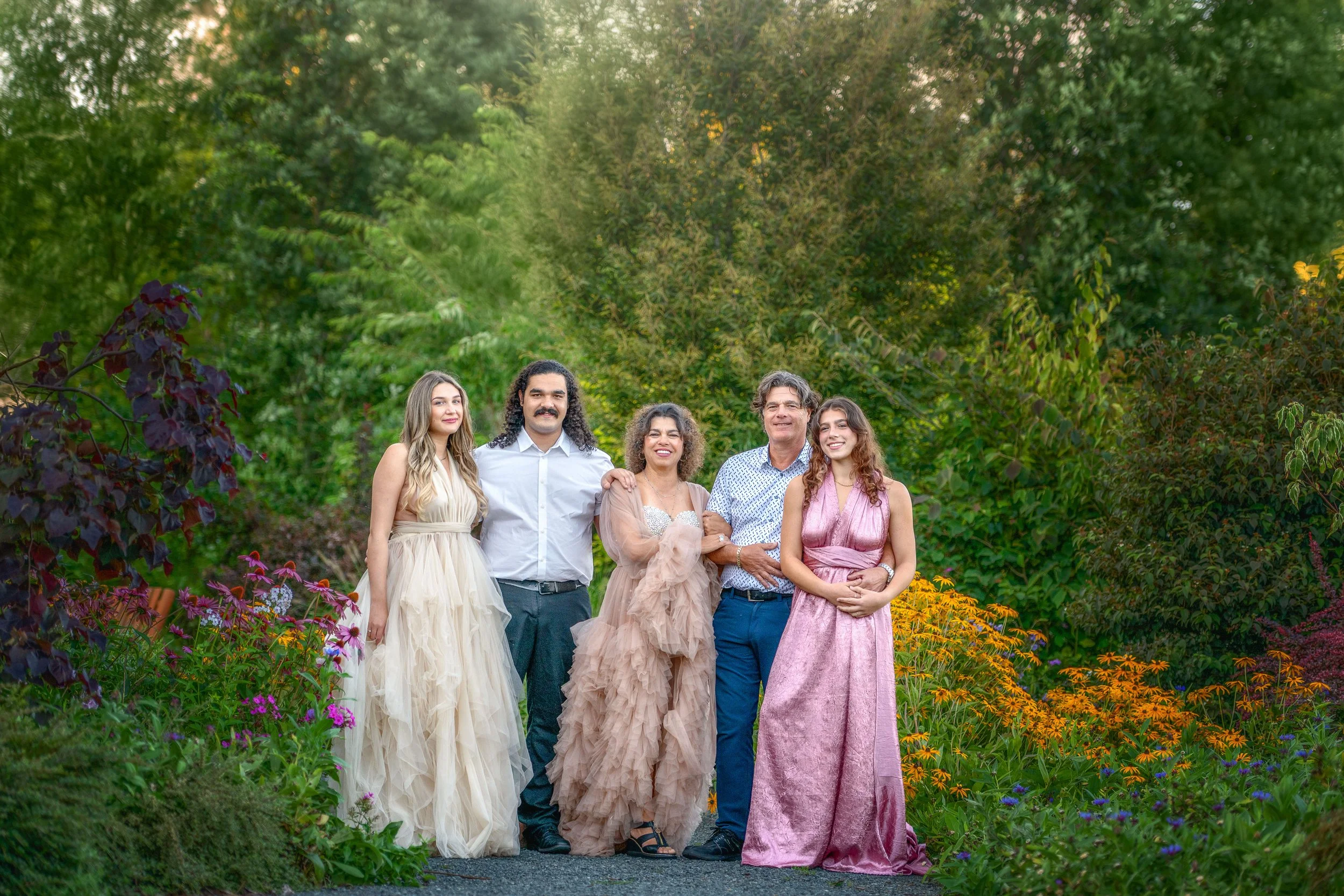 Group of five people dressed in formal attire standing outdoors among colorful flowers and green trees.