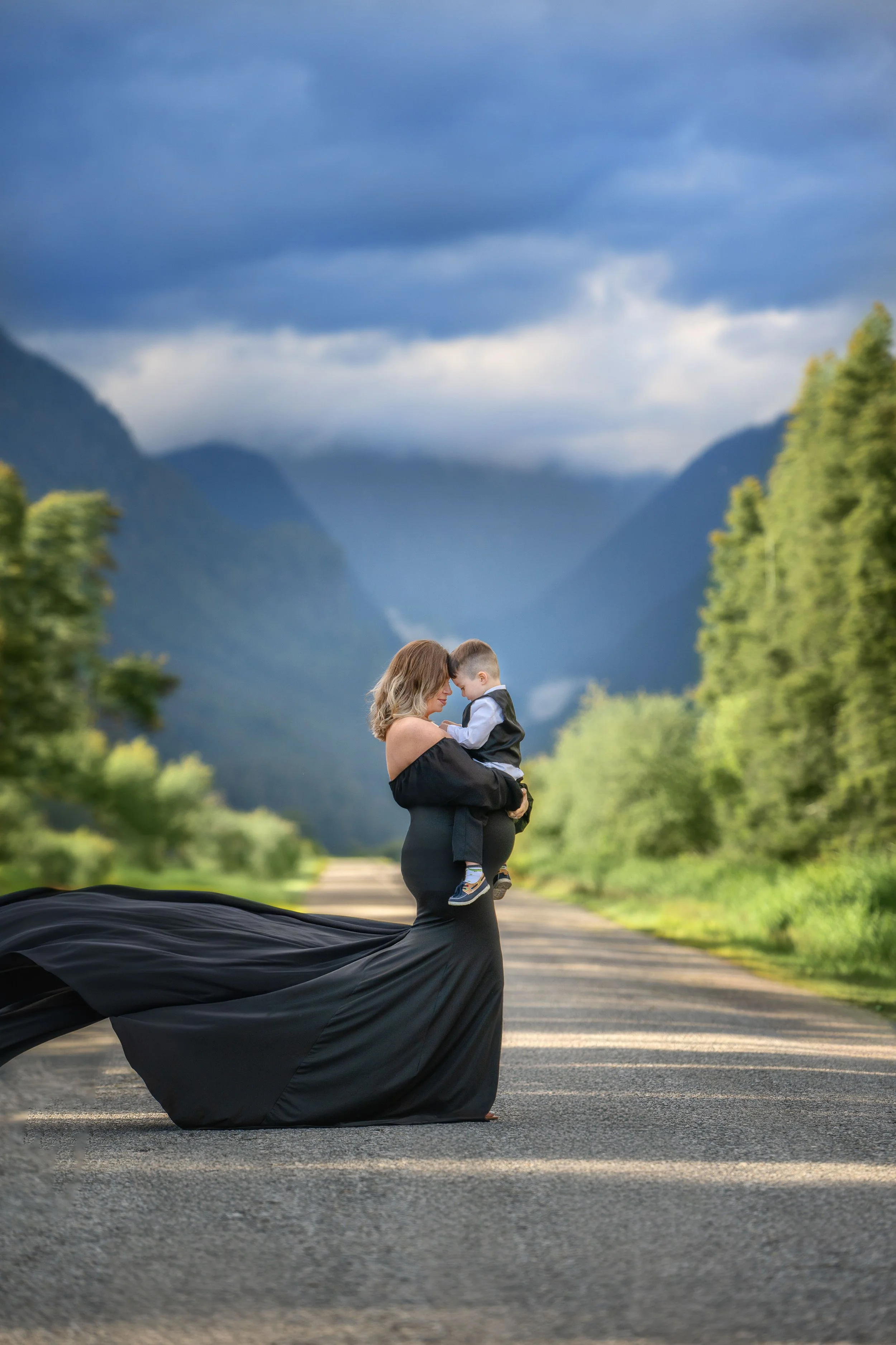 A woman in a black dress holding a young boy in a scenic outdoor setting with lush green trees and mountains in the background.