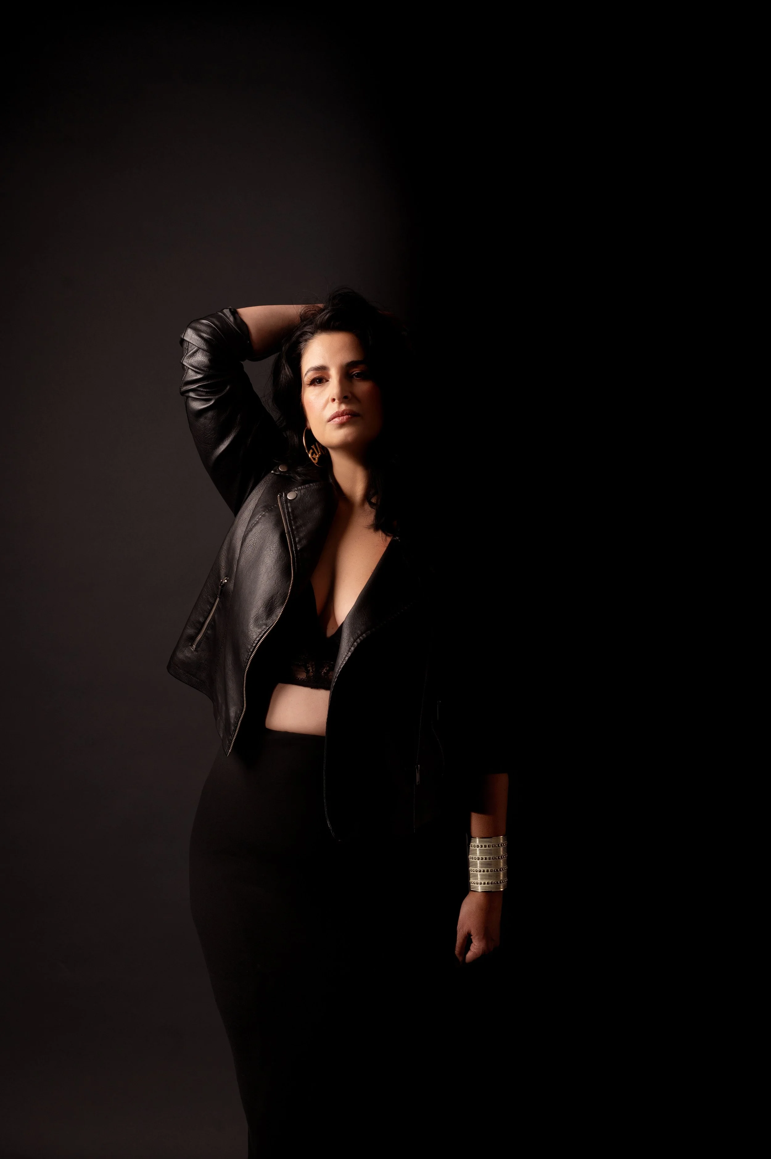 A woman with dark hair wearing a black leather jacket, black top, and a long black skirt, posing against a dark background with one arm raised behind her head.