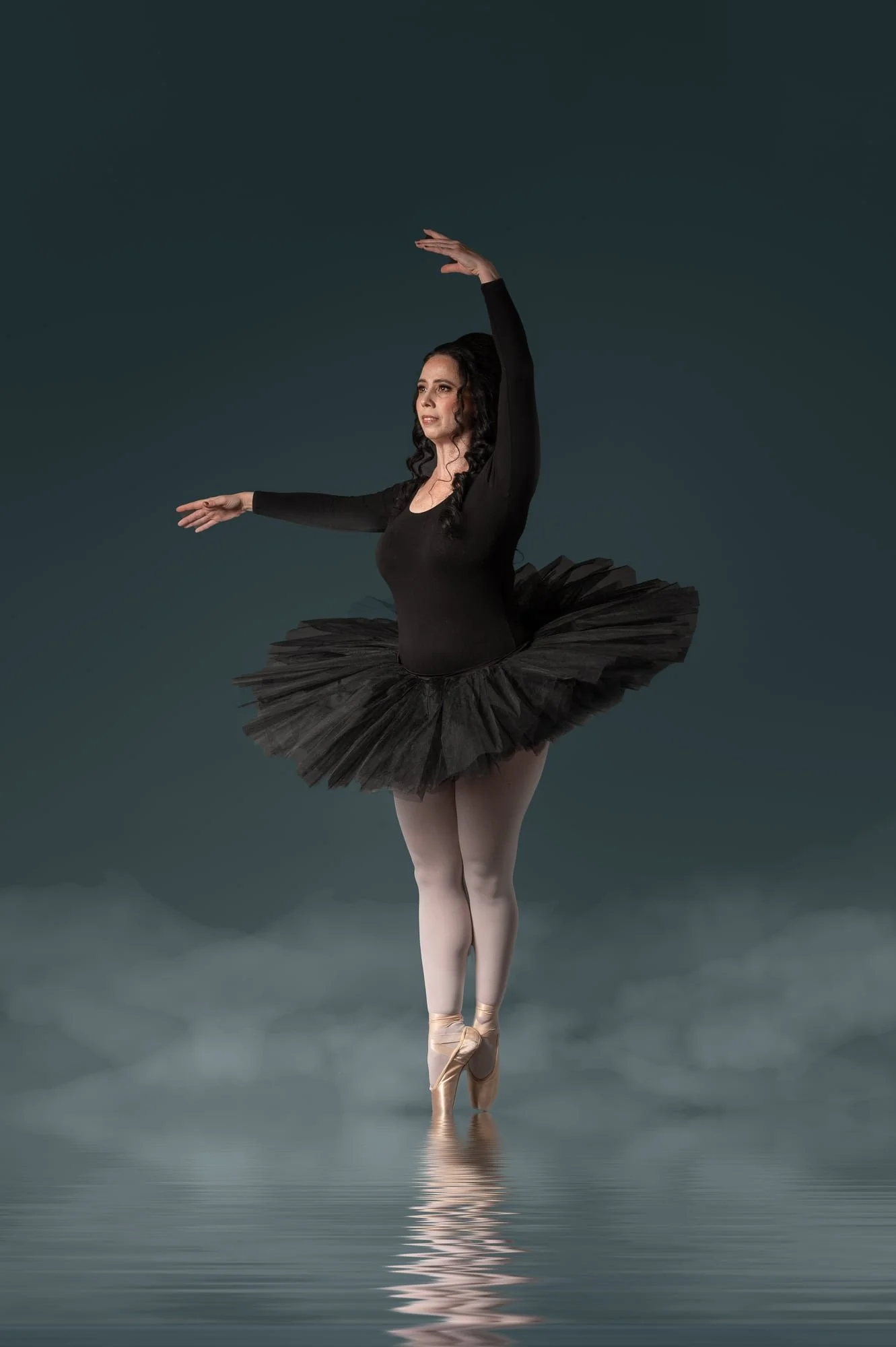 A ballerina performing on pointe in a black tutu, standing on water with a dark background.