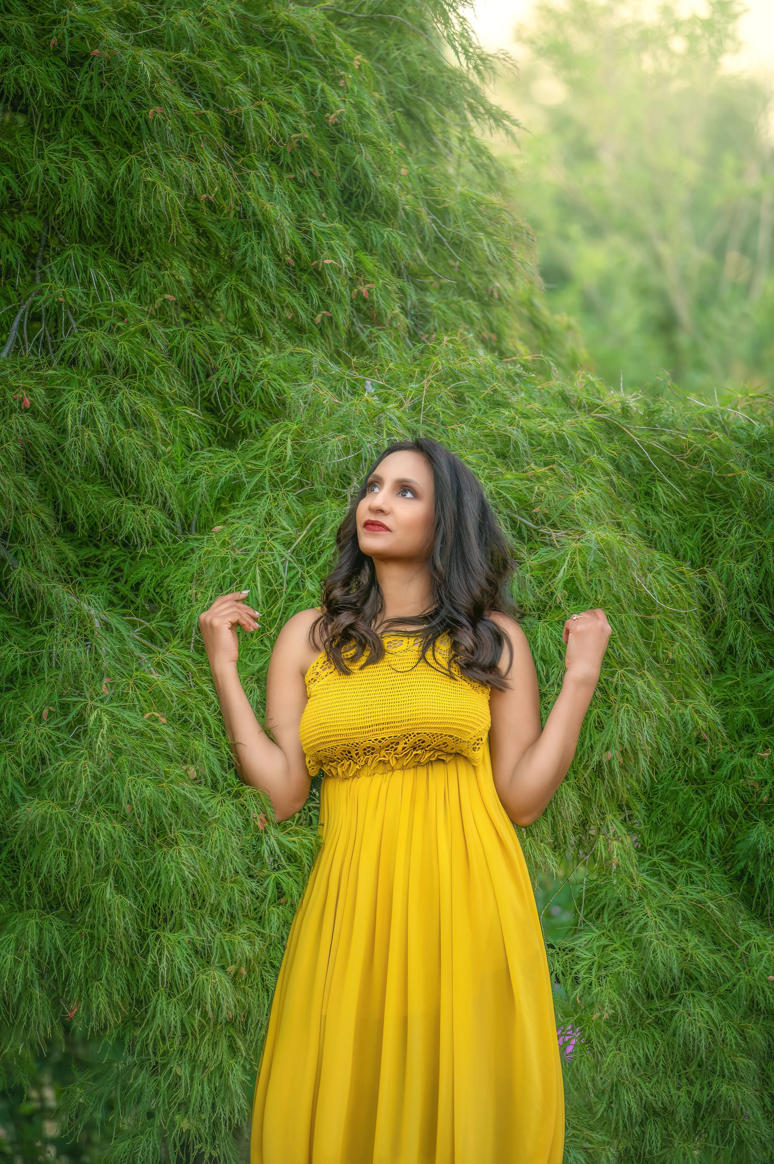 A woman in a yellow dress standing in front of lush green foliage, looking upward with her arms slightly raised.