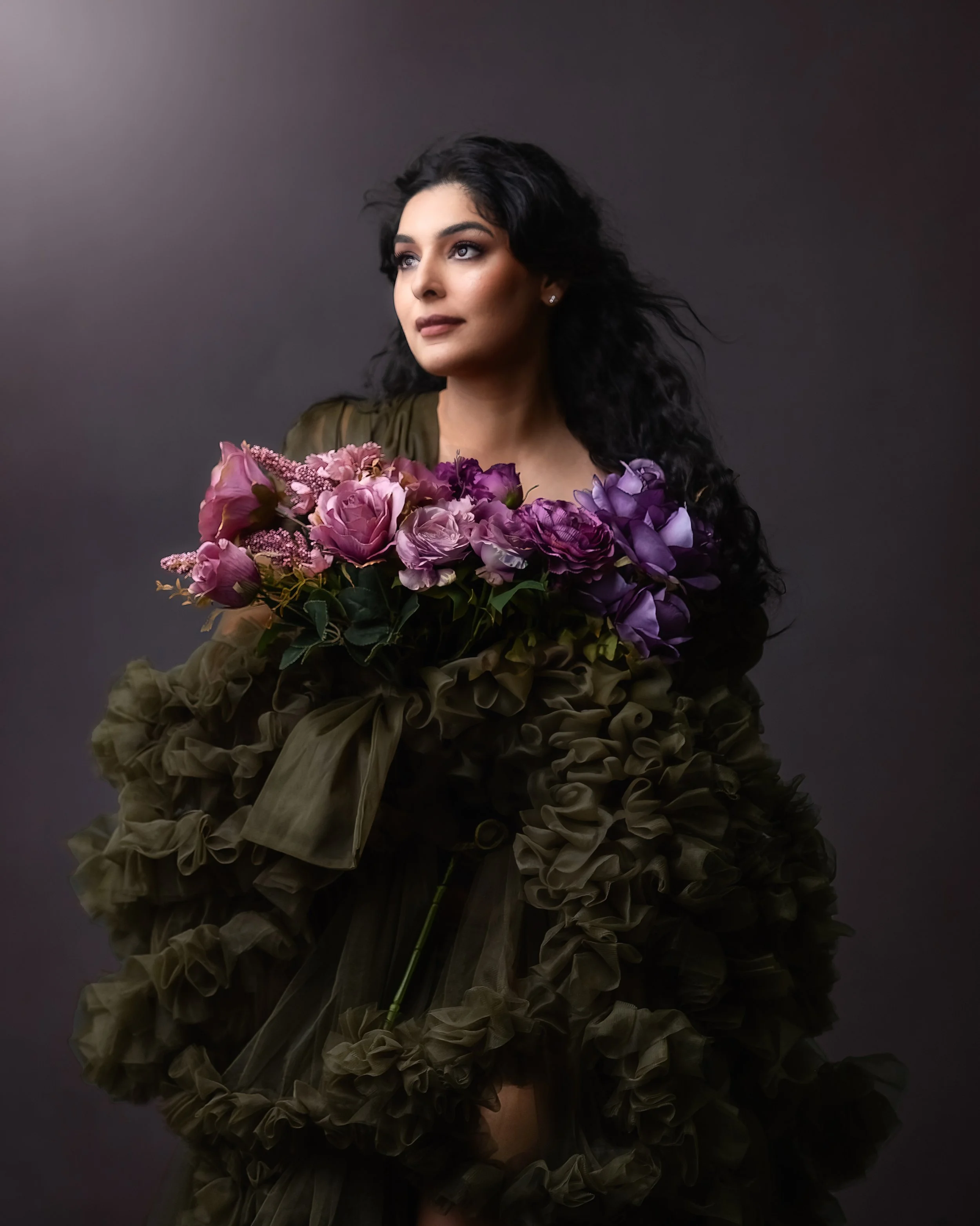 A woman with dark, curly hair holds a large bouquet of pink and purple flowers against a dark background.