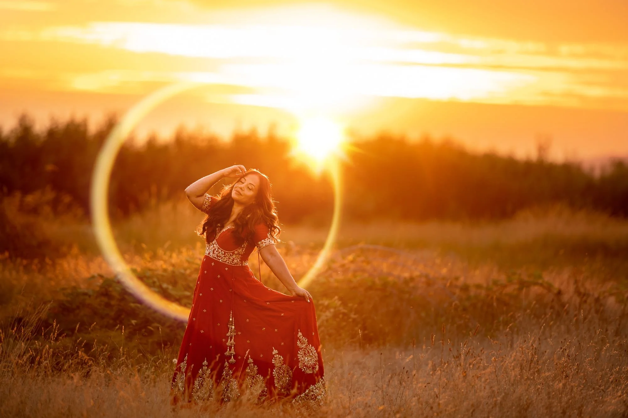 A woman in a red, embroidered dress standing in a field during sunset with the sun behind her, creating lens flare.