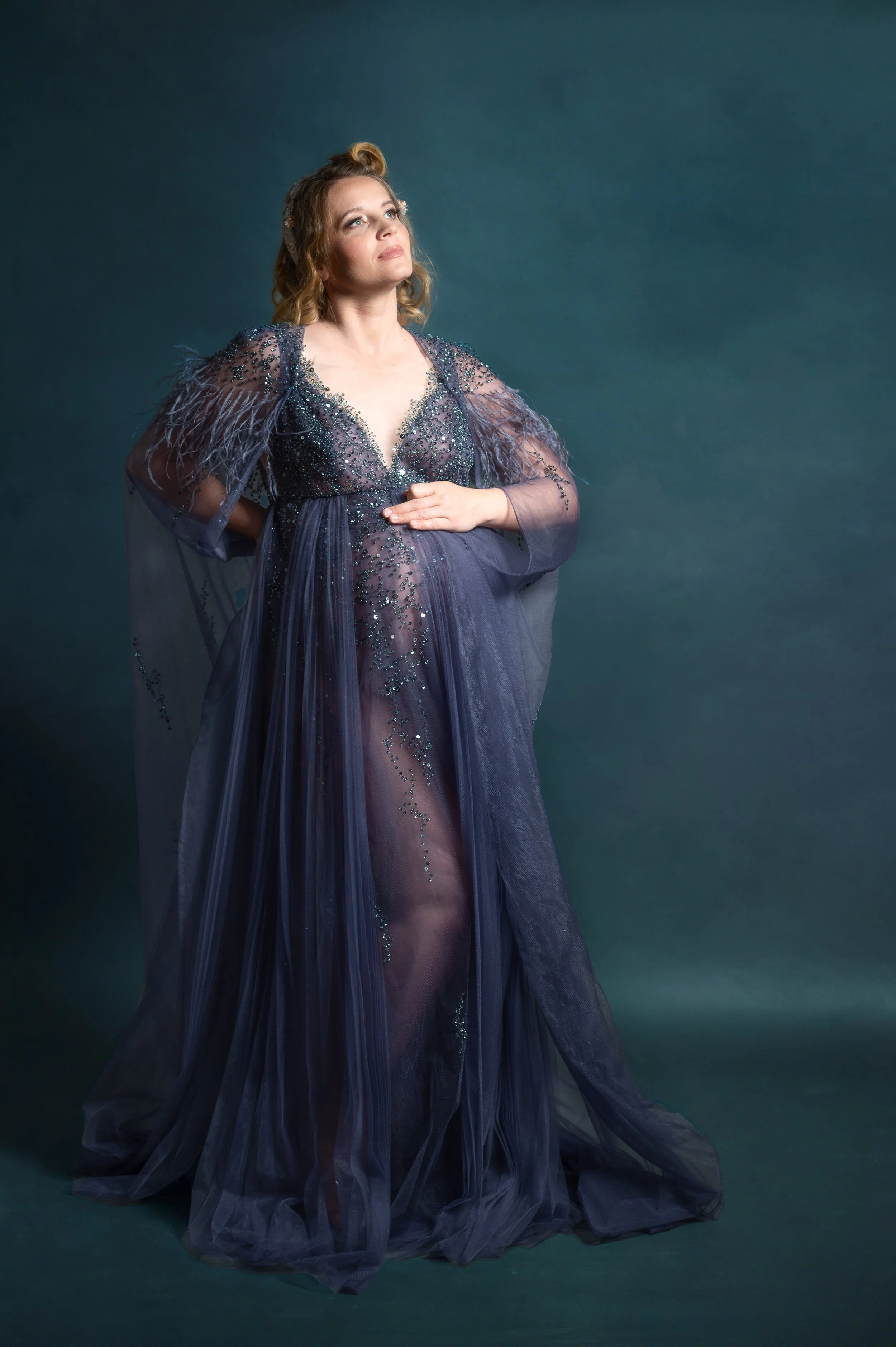 A pregnant woman wearing a sheer, decorated purple gown, posing against a dark background, with her hand on her belly and looking stylish and confident.