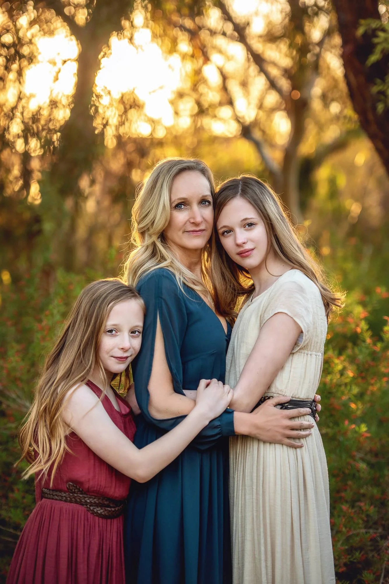 A woman with two girls poses in a park during sunset, surrounded by trees and greenery.