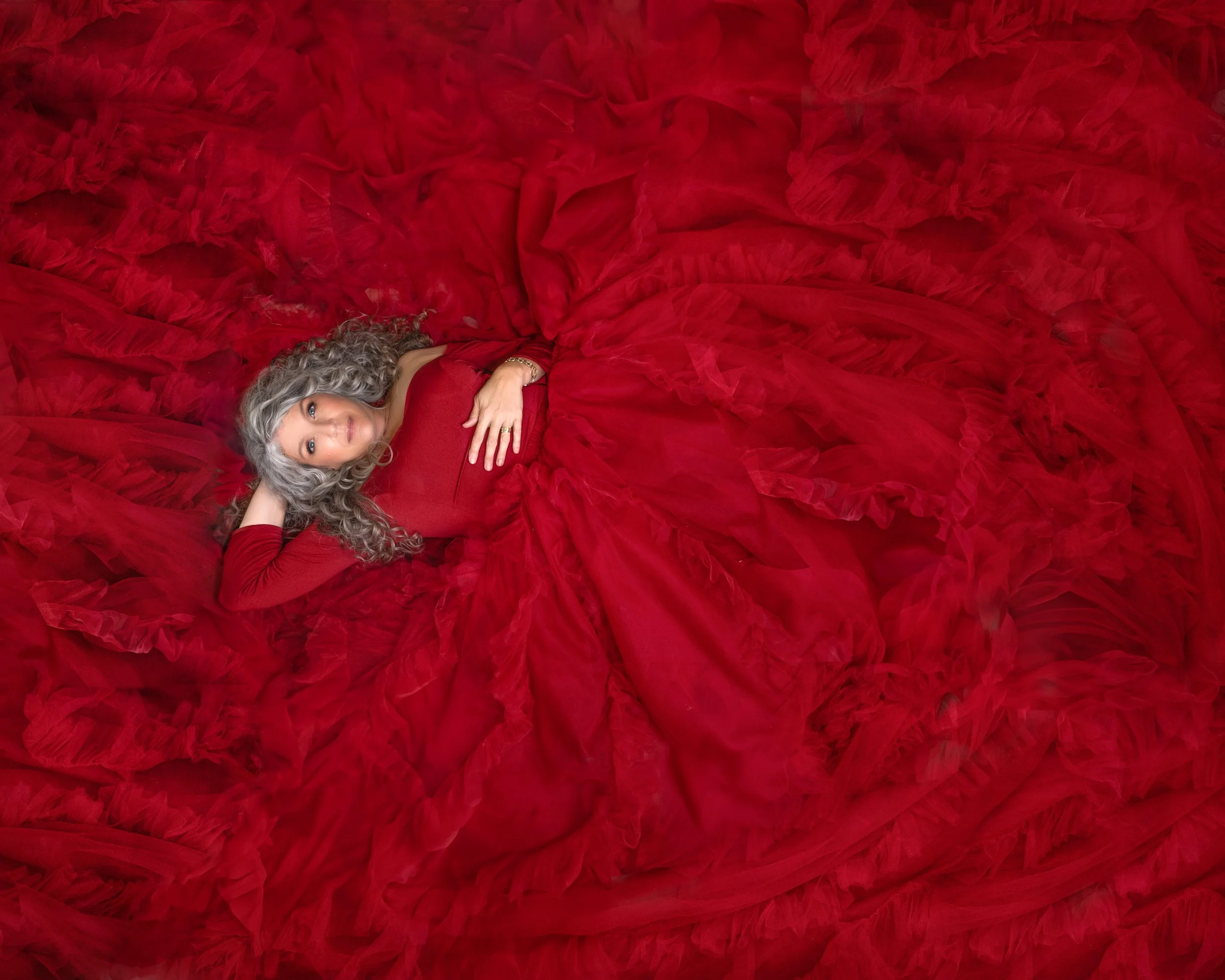 Woman in a red gown lying on her back with her hand on her stomach and her other arm behind her head, surrounded by ruffled red fabric.