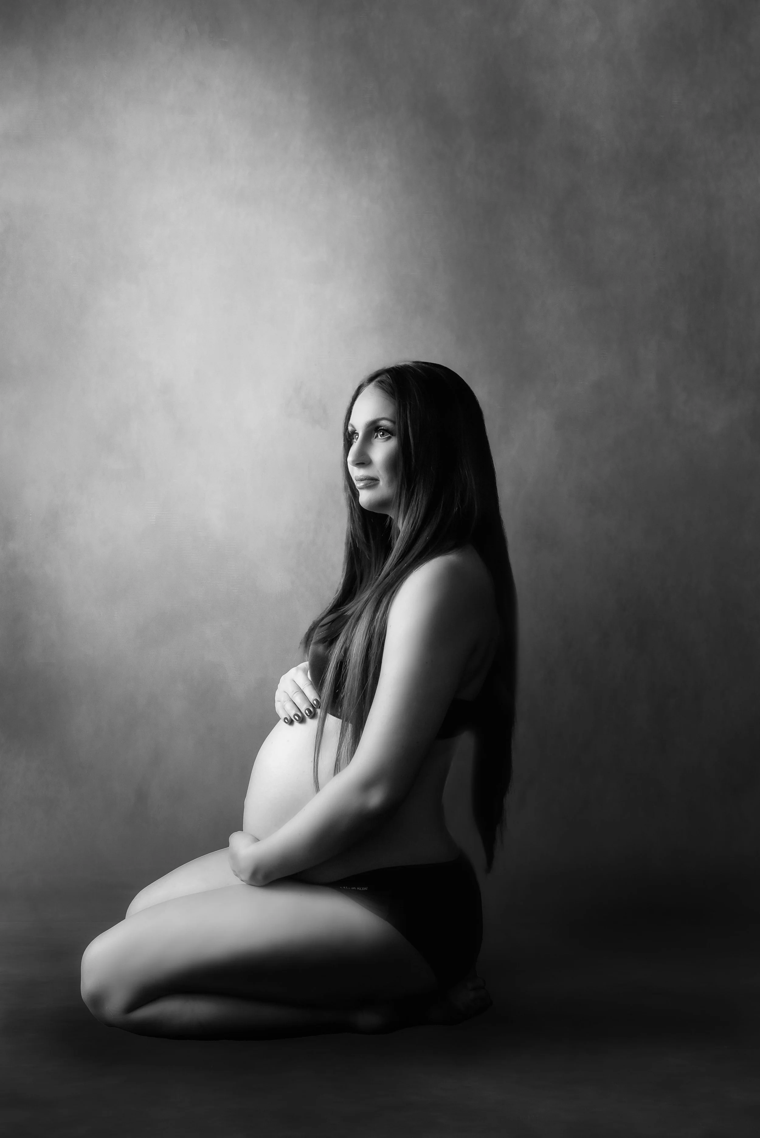 Black and white portrait of a pregnant woman sitting on the floor, holding her belly, with long hair, looking to the side against a plain background.