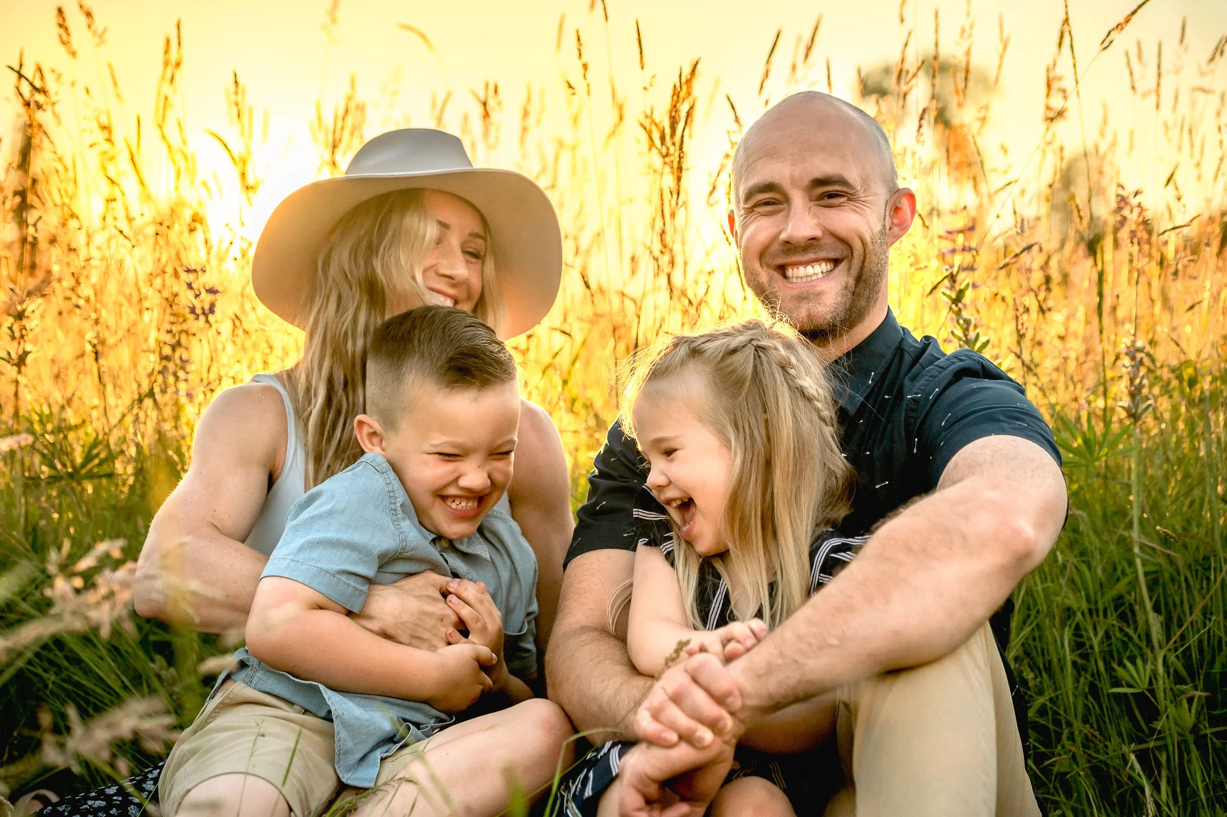 A family of four sitting on grass in a field during sunset, smiling and joking around. The parents are a man and a woman, with the woman wearing a large white hat. Their two children, a boy and a girl, are laughing and hugging each other.
