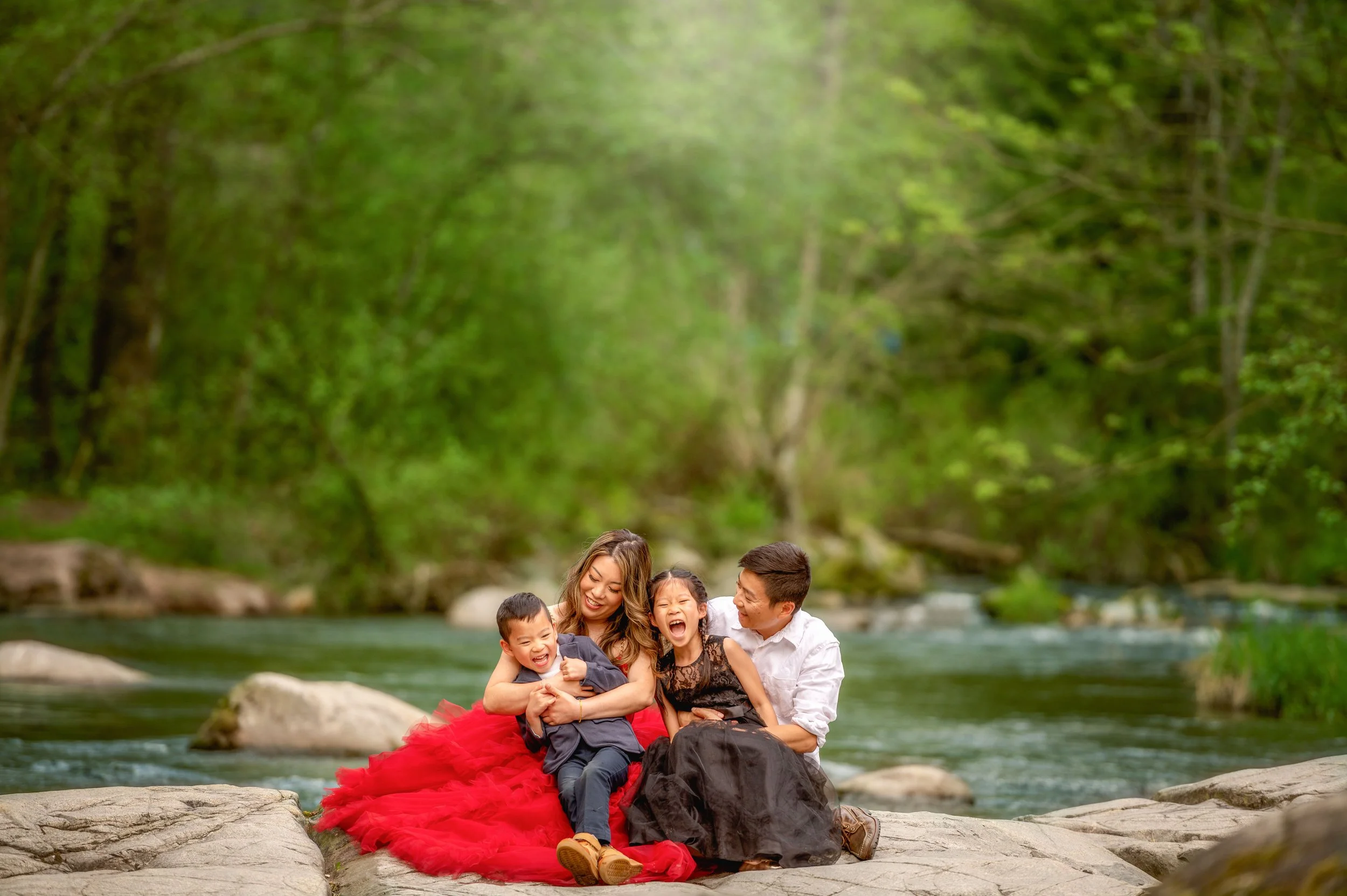 A family of four enjoying time by a river surrounded by lush green trees, sitting on rocks with happy expressions.
