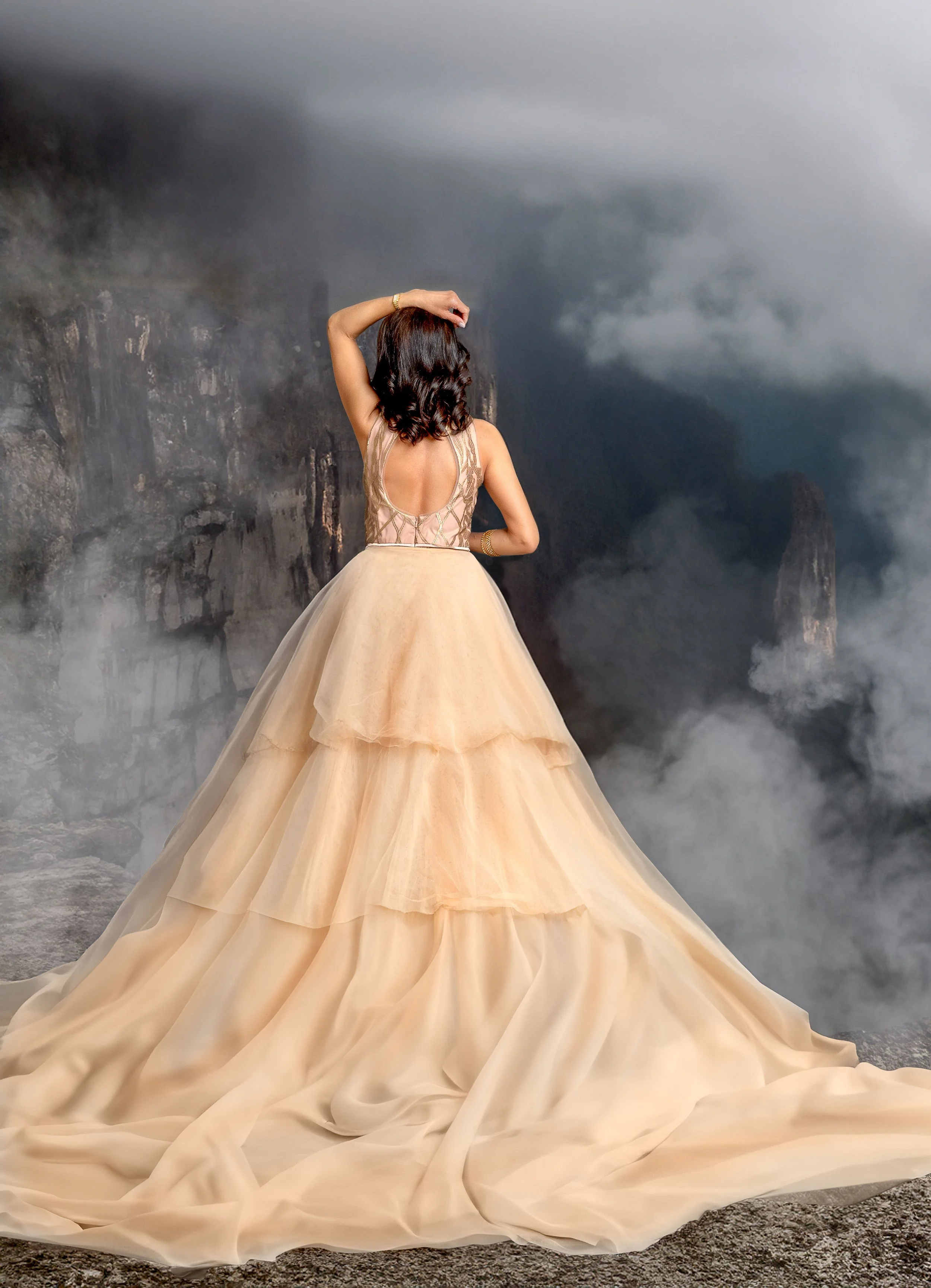 Woman in an elegant, flowing beige gown standing on rocky terrain with a misty mountain backdrop.
