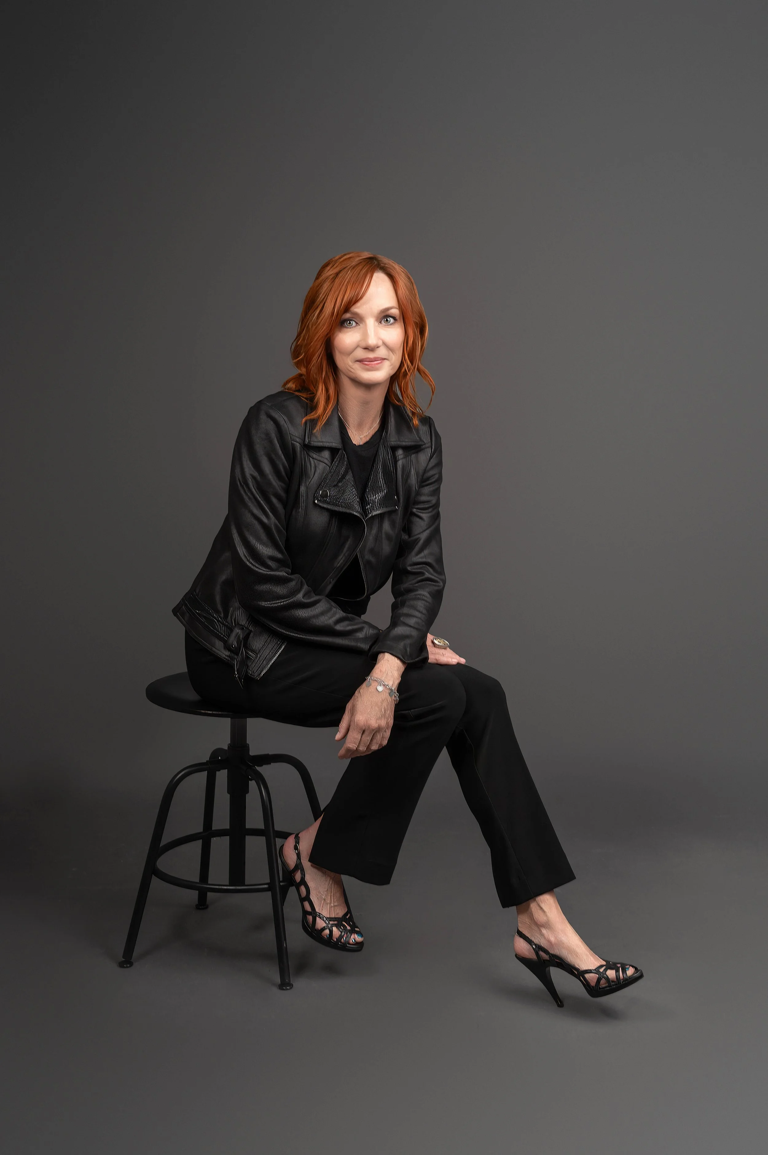 A woman with red hair wearing a black leather jacket, black pants, and black high-heeled shoes sitting on a stool against a gray background.