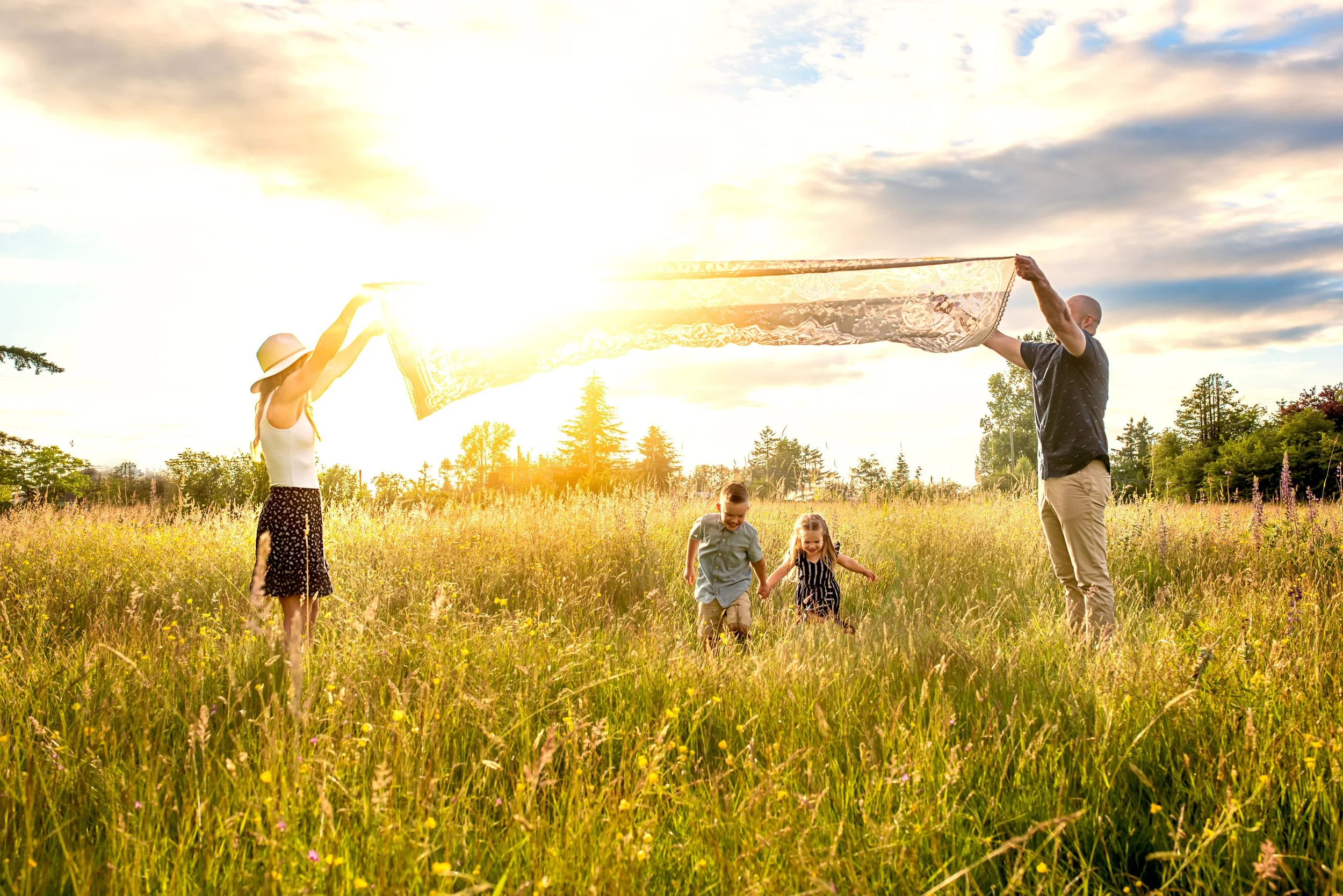 A family of four playing in a grassy field during sunset, with two adults holding a blanket and two children running through the grass.