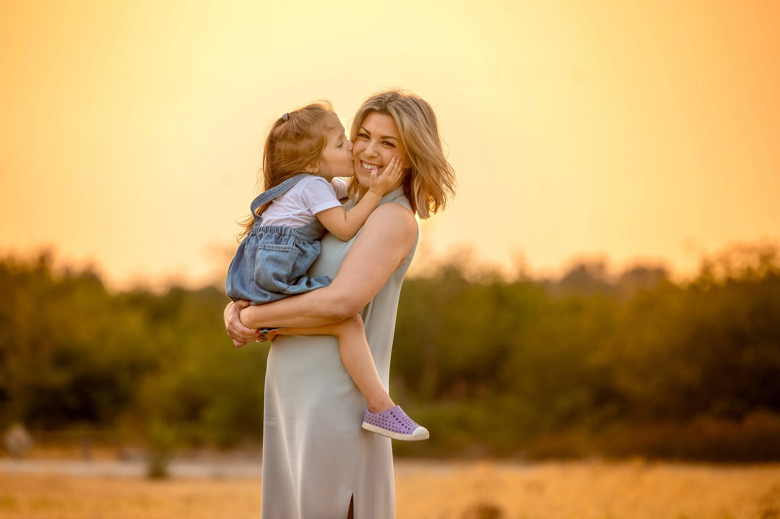 A woman holding a young girl outdoors at sunset, with the girl giving the woman a kiss on the cheek.