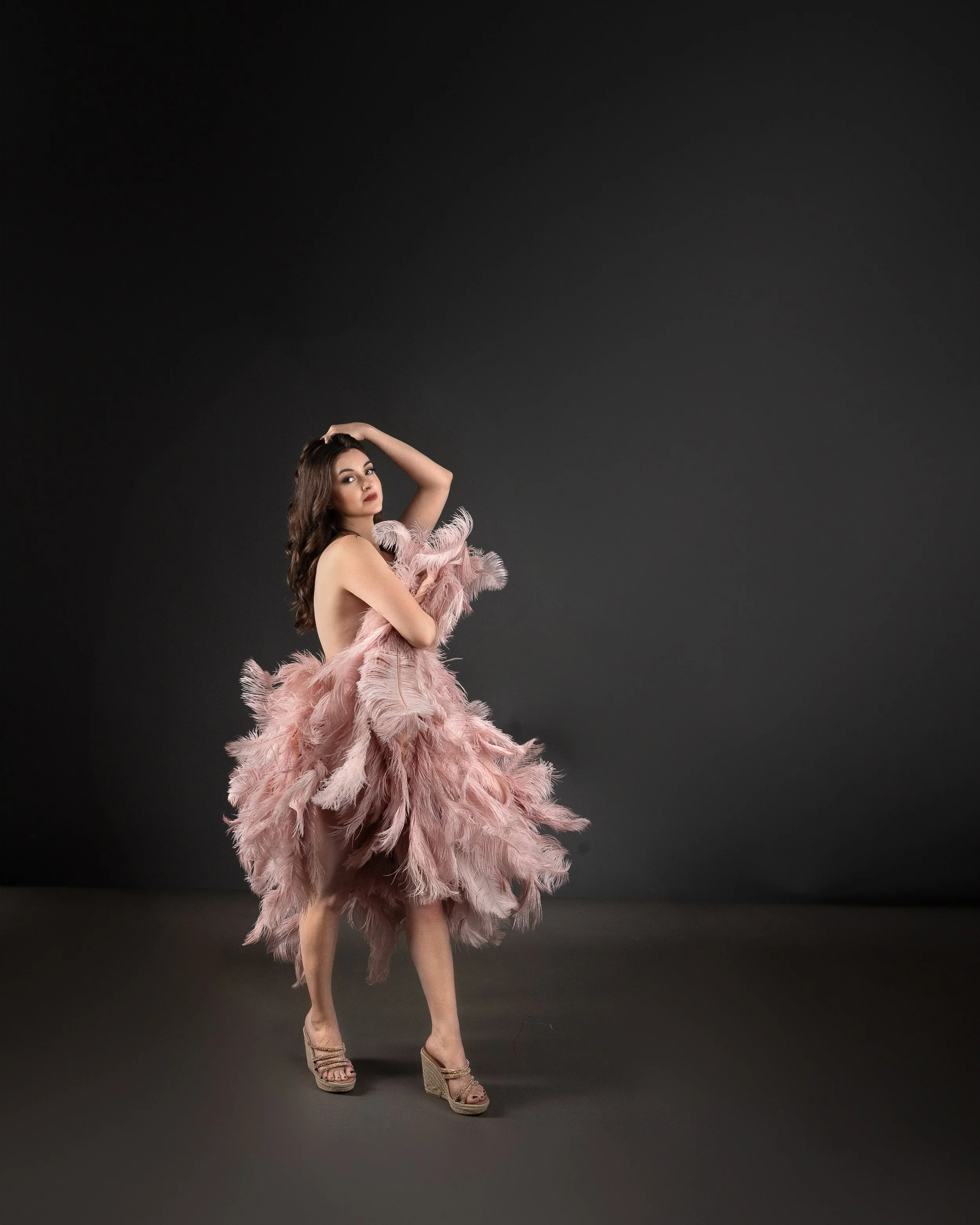 A woman stands against a dark background, wearing spiral, open-toed heels and a pink feathered dress, with her right arm raised and her left arm across her body.