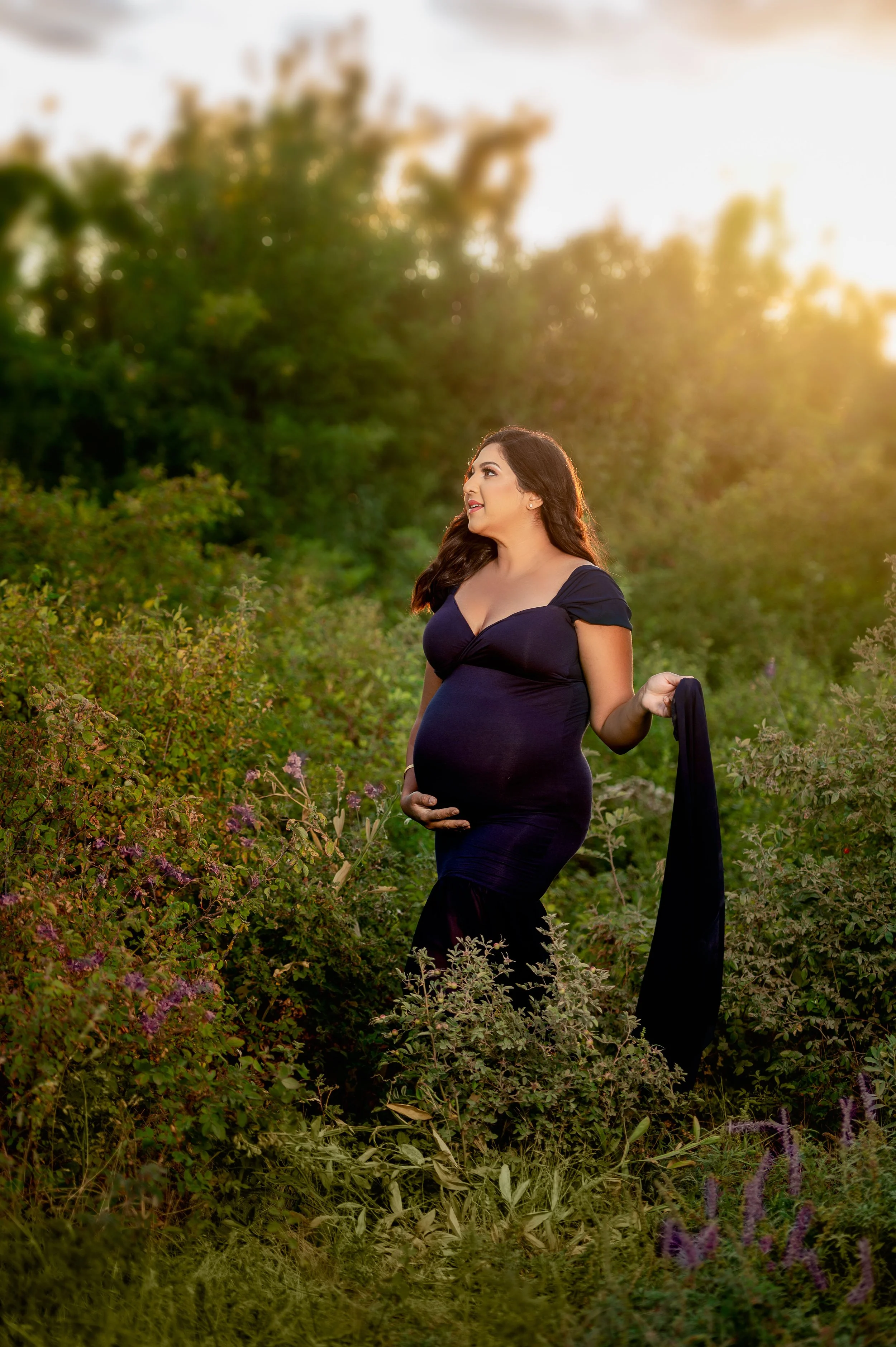 Pregnant woman in a dark dress standing among greenery with sunlight in the background.