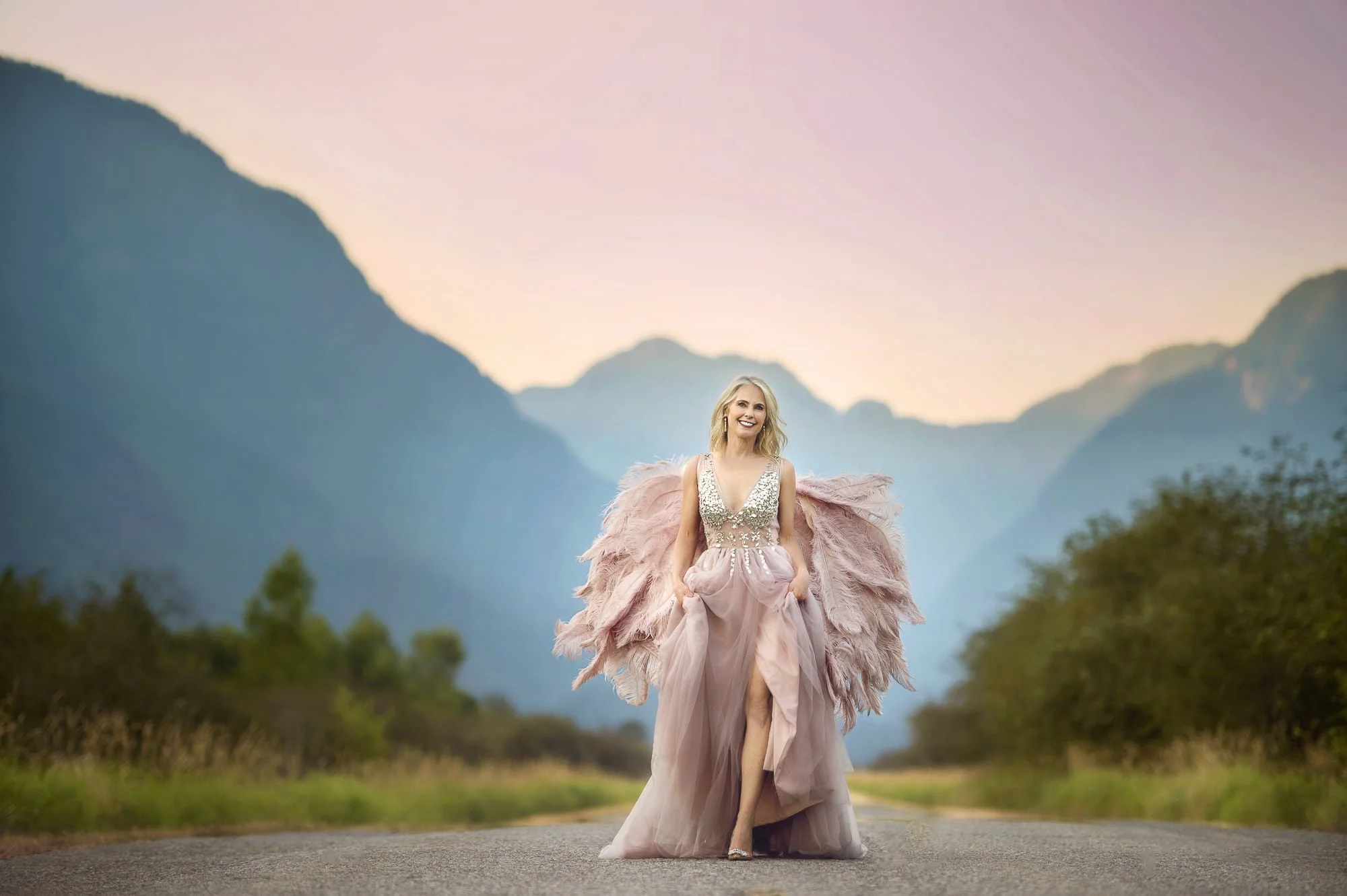Woman in a strapless gown with feather details walking on a rural road at sunset, mountains in the background.