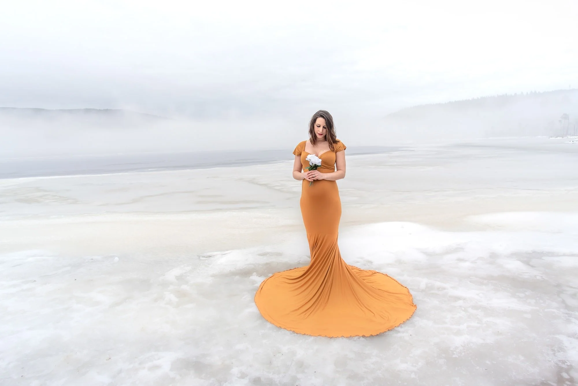 Woman in a long, orange dress standing on a snowy or icy landscape holding white flowers, with foggy background and distant hills.