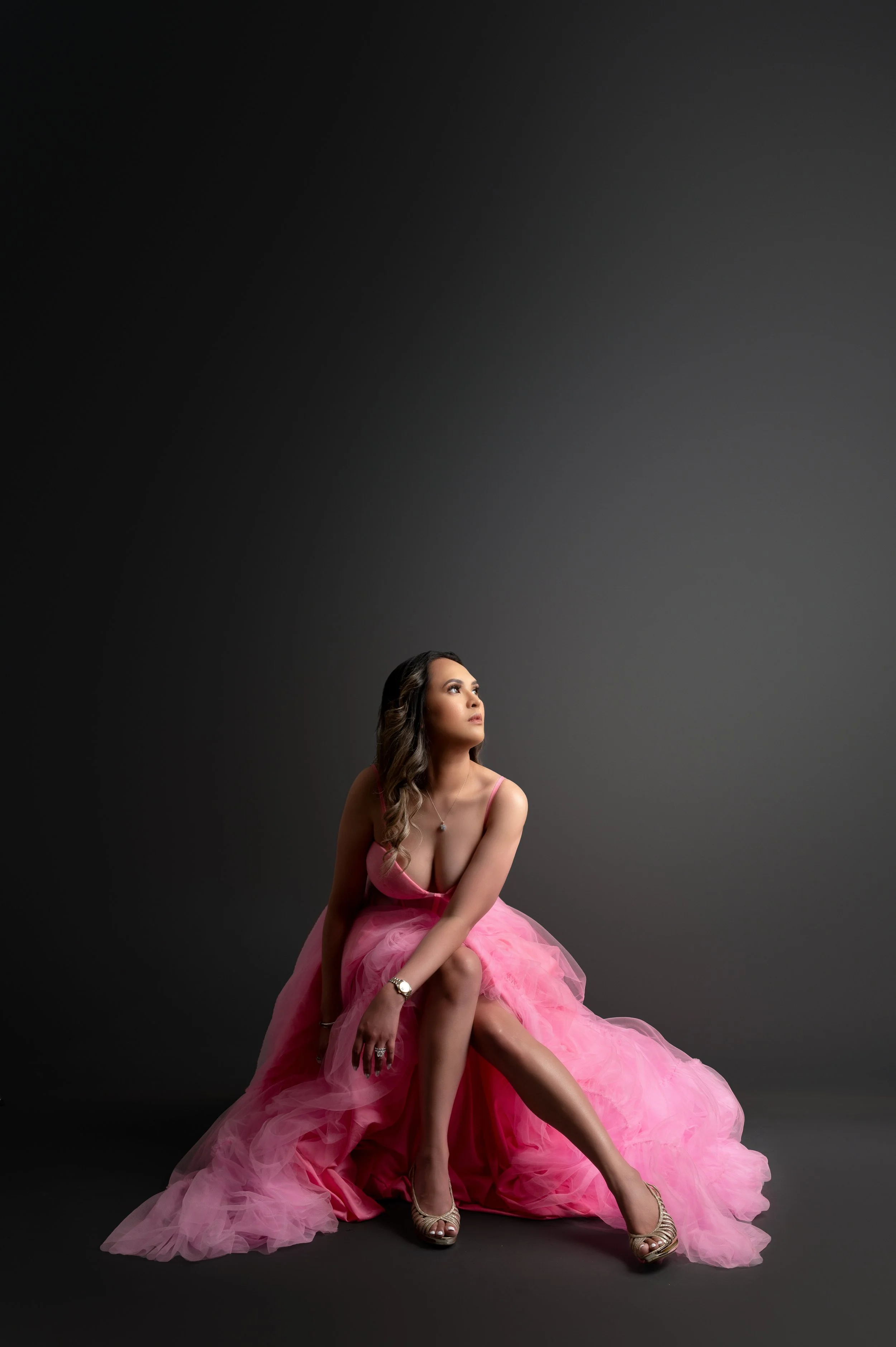 A woman in a pink gown sitting on the floor against a dark background, looking upwards.
