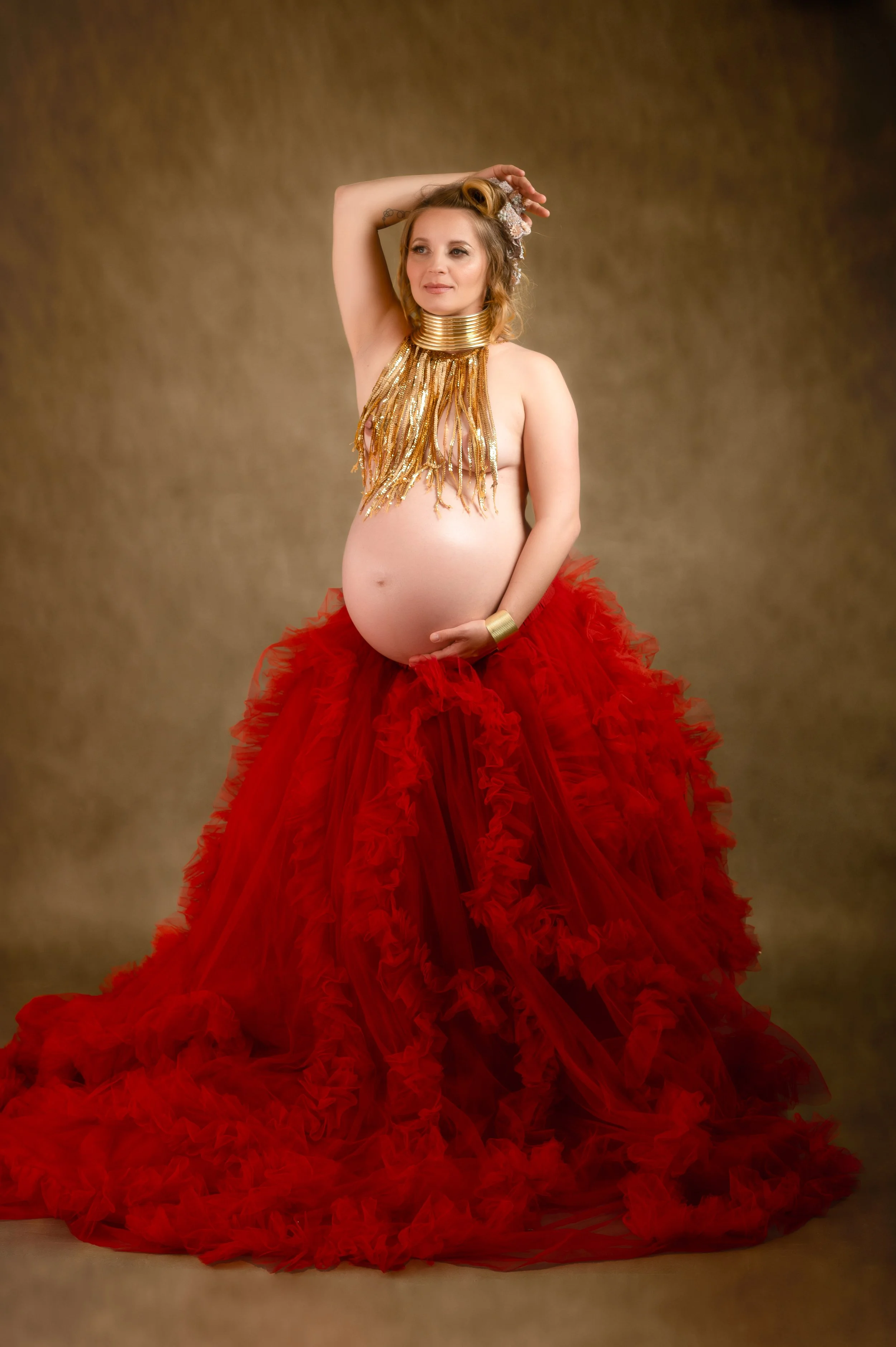 Pregnant woman wearing a gold fringed necklace, gold cuff bracelet, and red ruffled skirt against a neutral background.